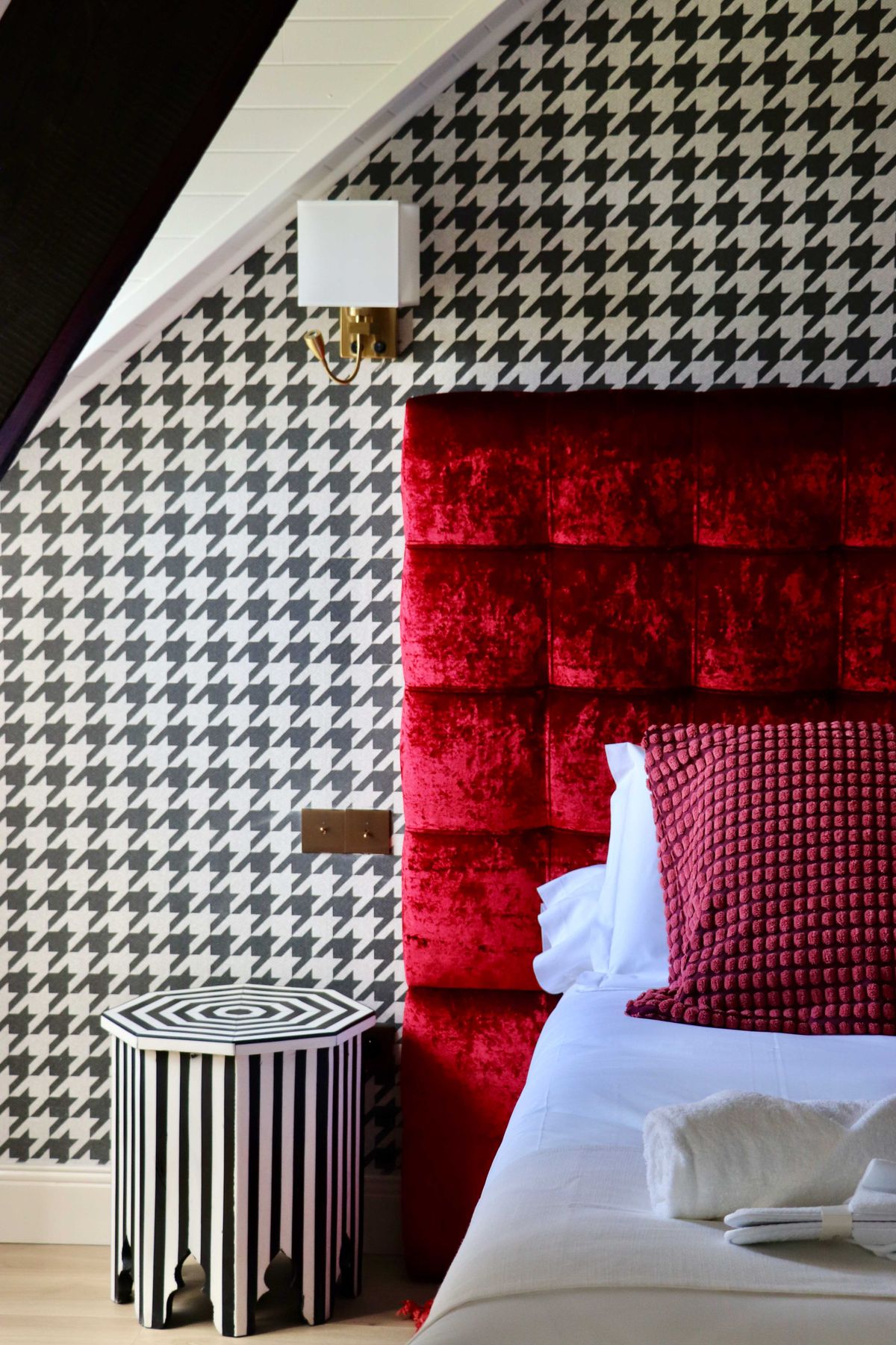 Boutique bedroom with houndstooth wallpaper, red velvet tufted headboard, and striped octagonal side table