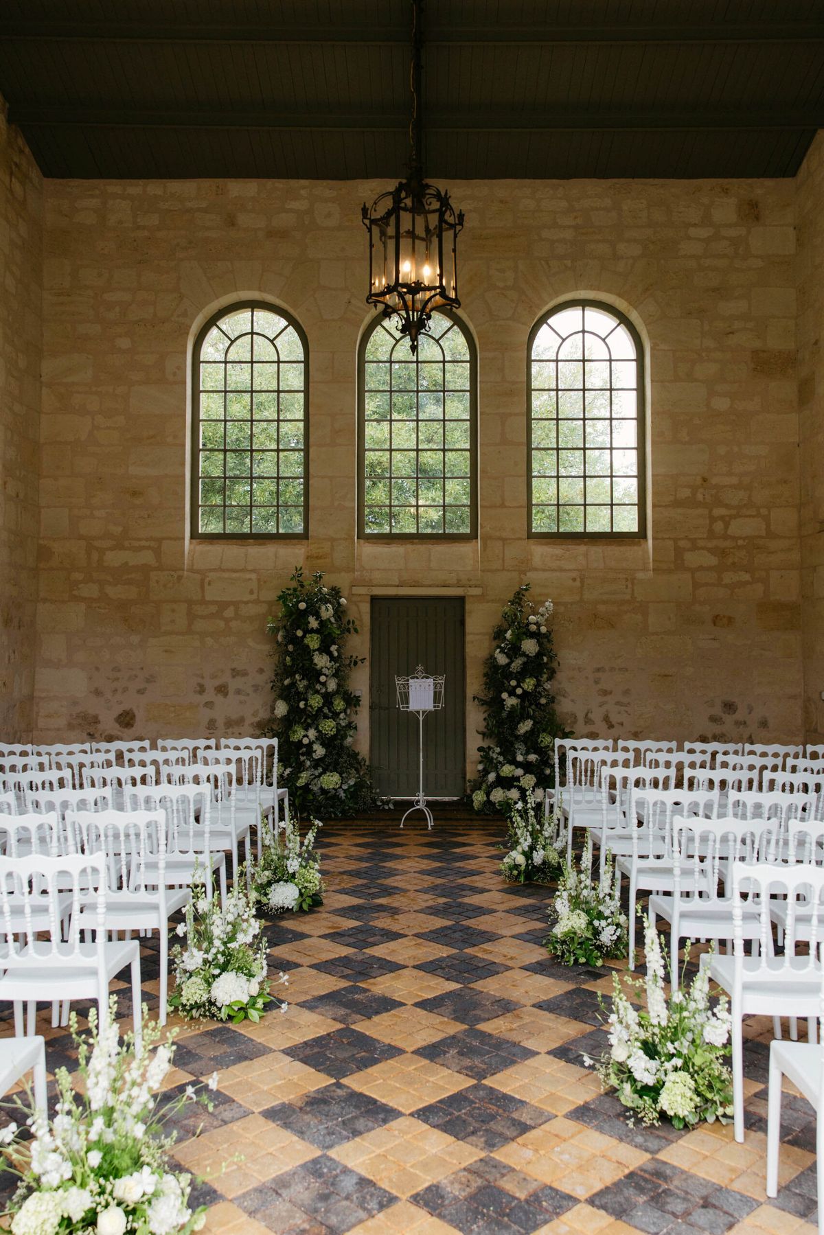 White chiavari chairs and hydrangea column arrangements in limestone chapel with checkerboard tile floor