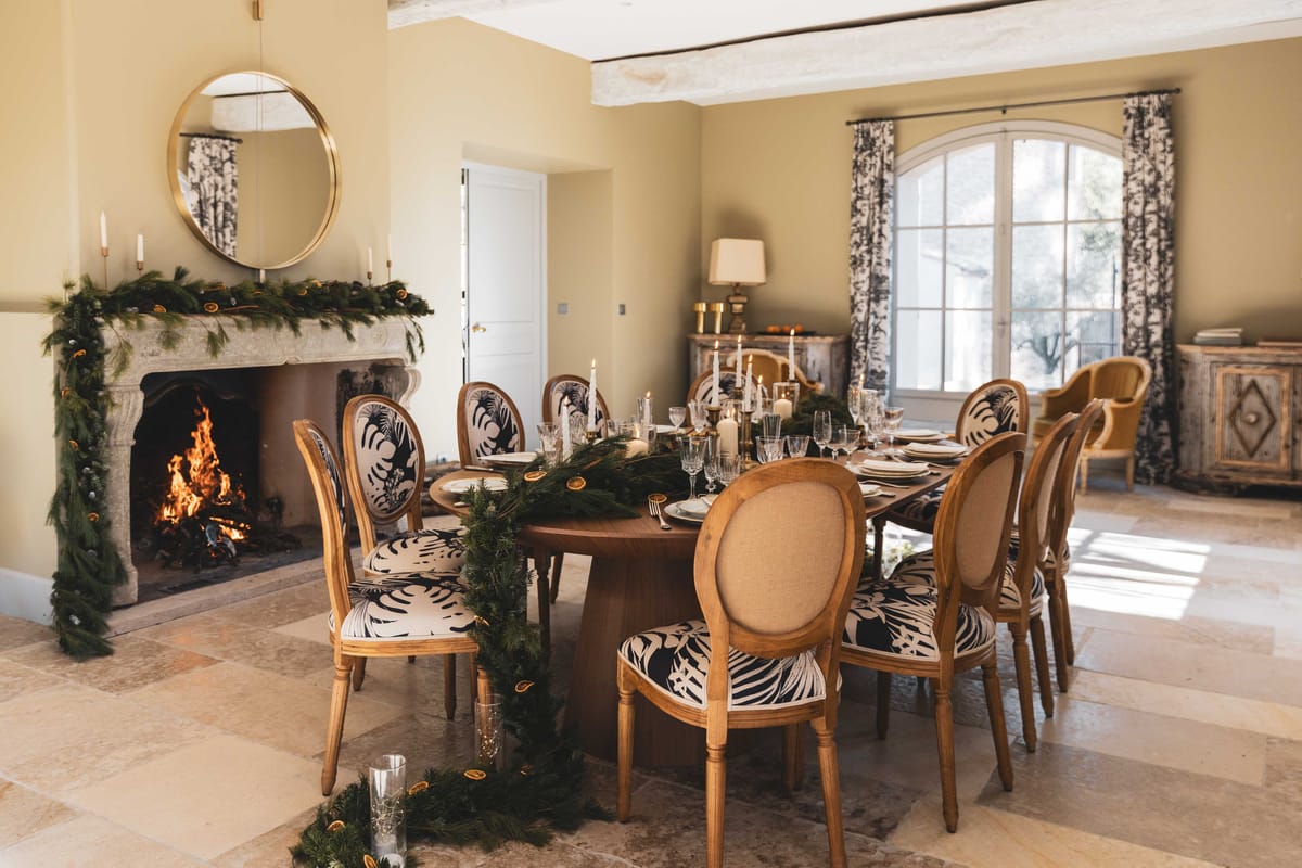 Winter dining room with pine and dried citrus garland, active fireplace, navy leaf-print chairs, and gold mirror
