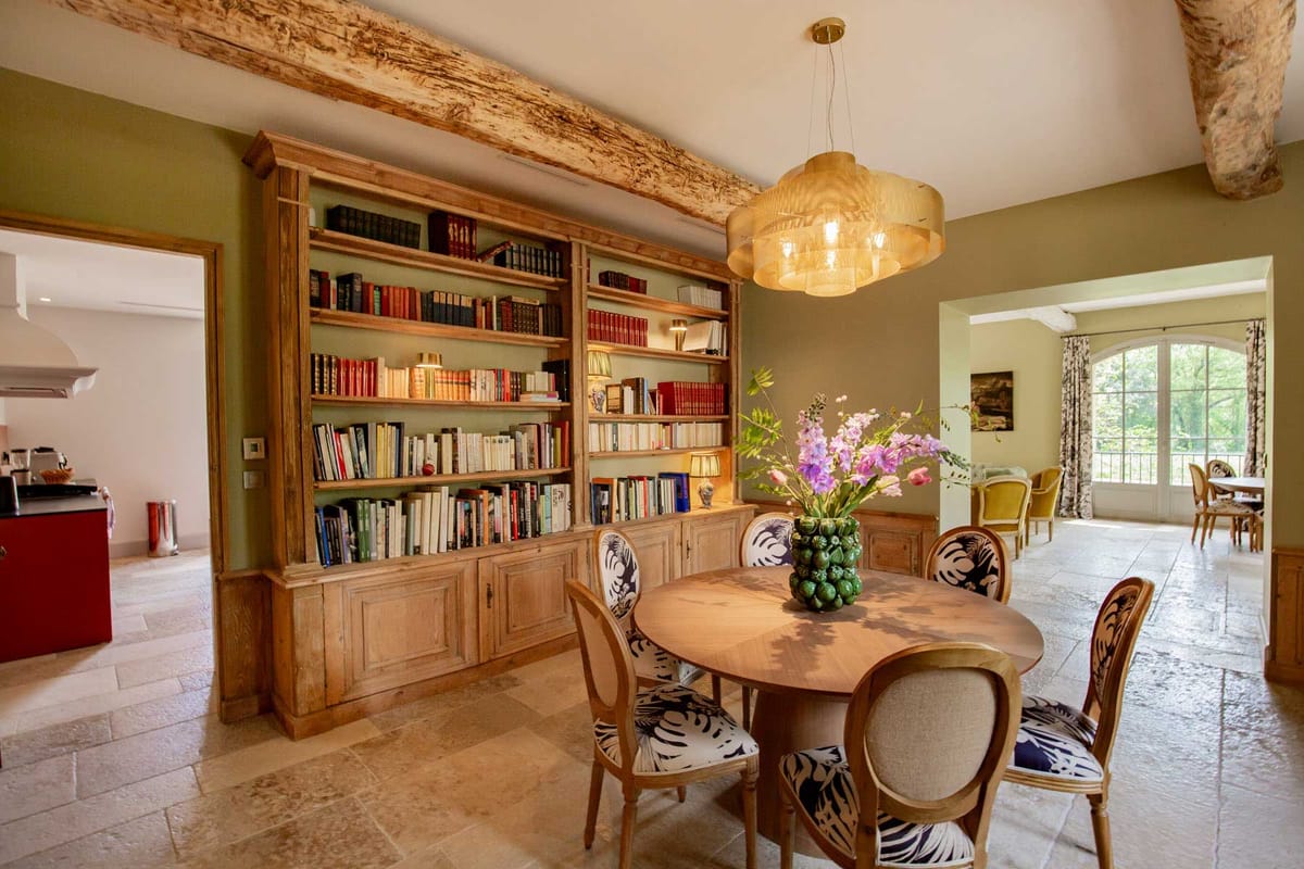 Dining room with exposed beams, olive walls, tropical-print chairs, oak bookcase, and gold pendant light
