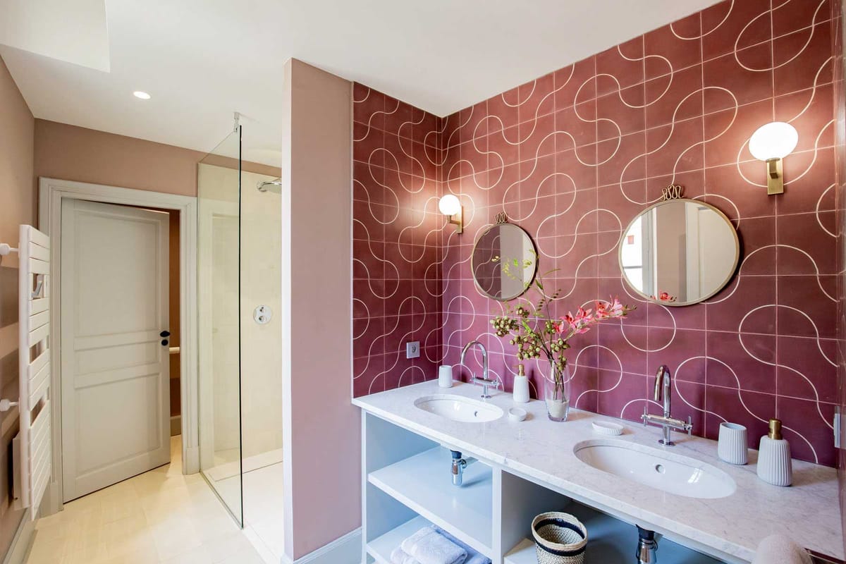 Modern bathroom with mauve patterned tiles, brass-framed mirrors, globe sconces, and pink orchid arrangement