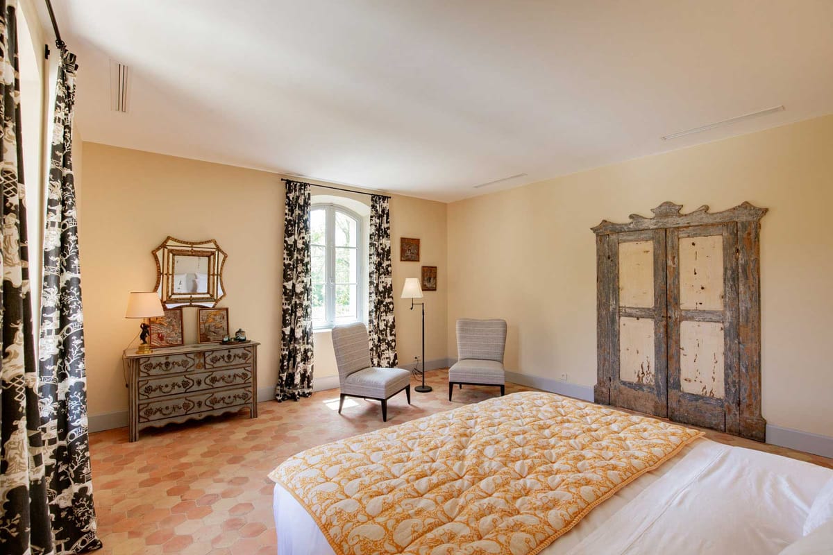 Chateau bedroom with peach walls, toile de Jouy curtains, distressed antique armoire, and hexagonal terracotta tiles
