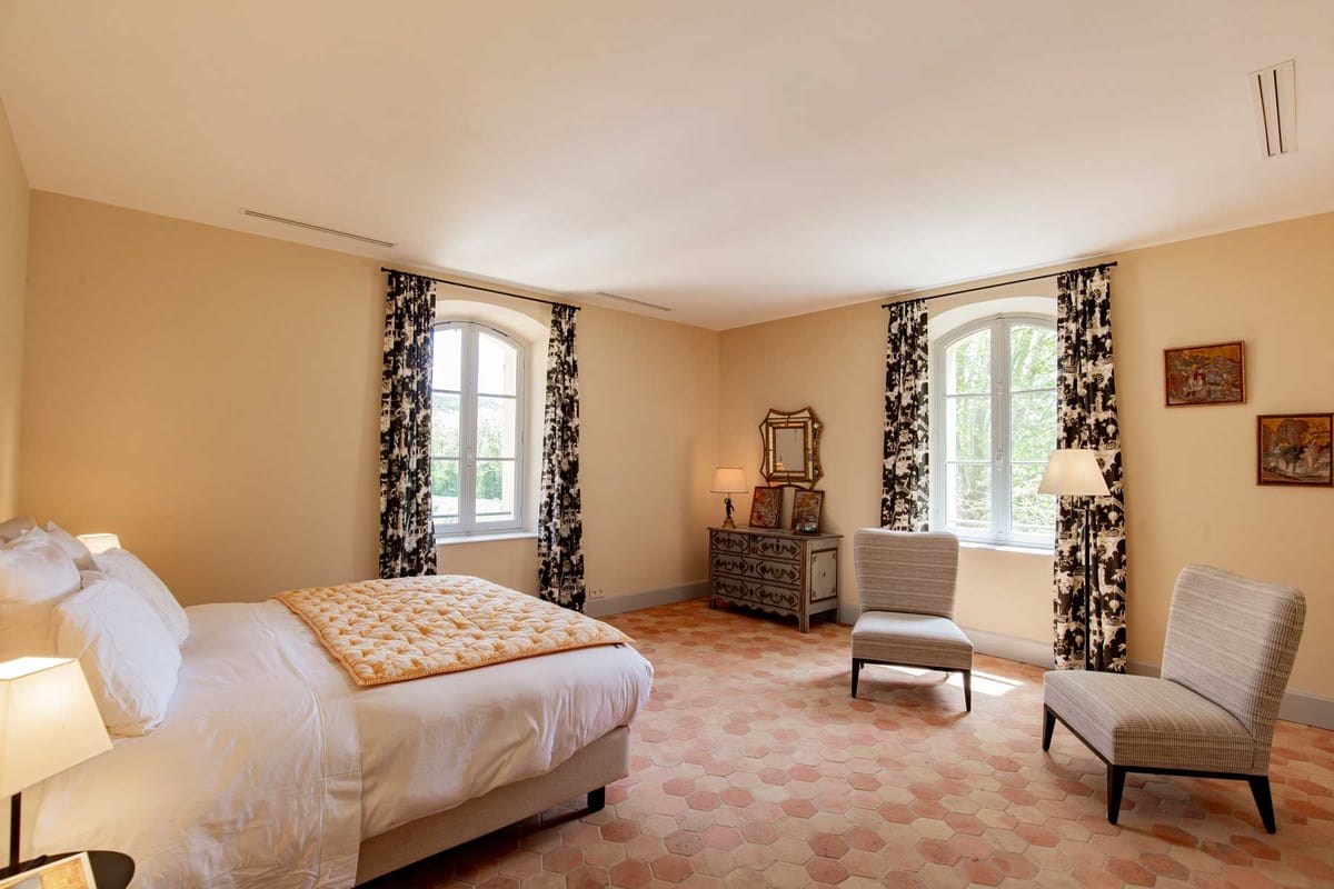 French country bedroom with terracotta tiles, toile curtains, white bed with gold throw, and grey armchairs