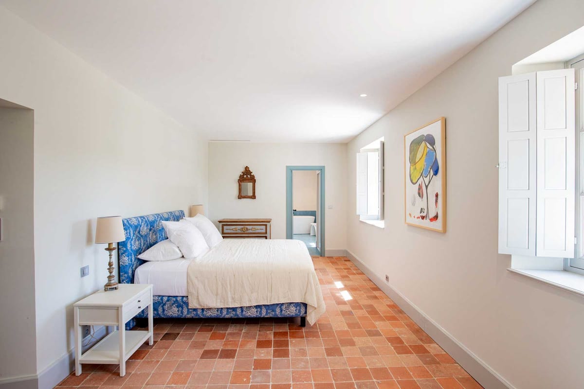Guest bedroom with blue toile headboard, terracotta tiles, abstract art, and freestanding bathtub through teal doorway