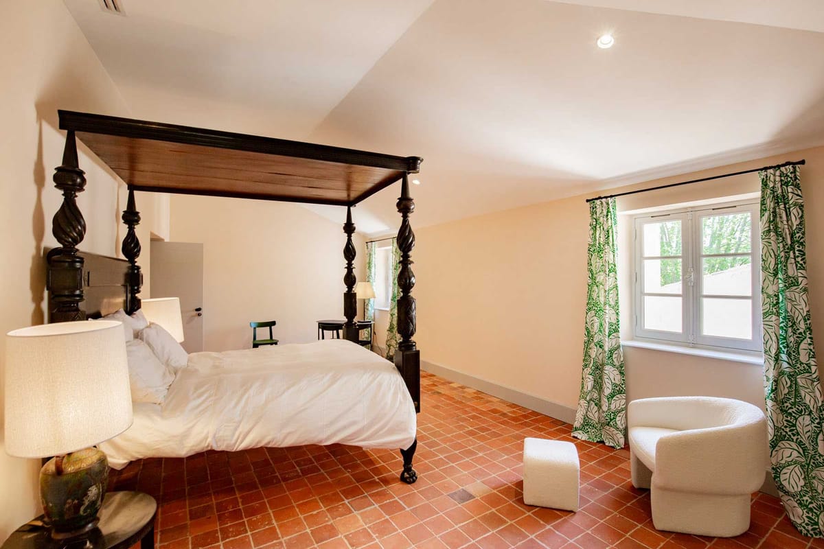 Guest room with carved four-poster bed, terracotta tiles, boucle armchair, and botanical curtains