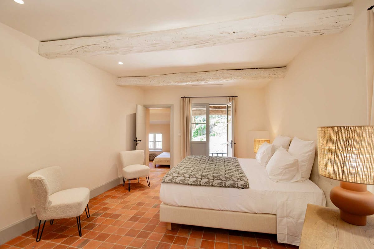 Provencal bedroom with whitewashed beams, terracotta floors, white linen bed, and sage armoire beside French doors