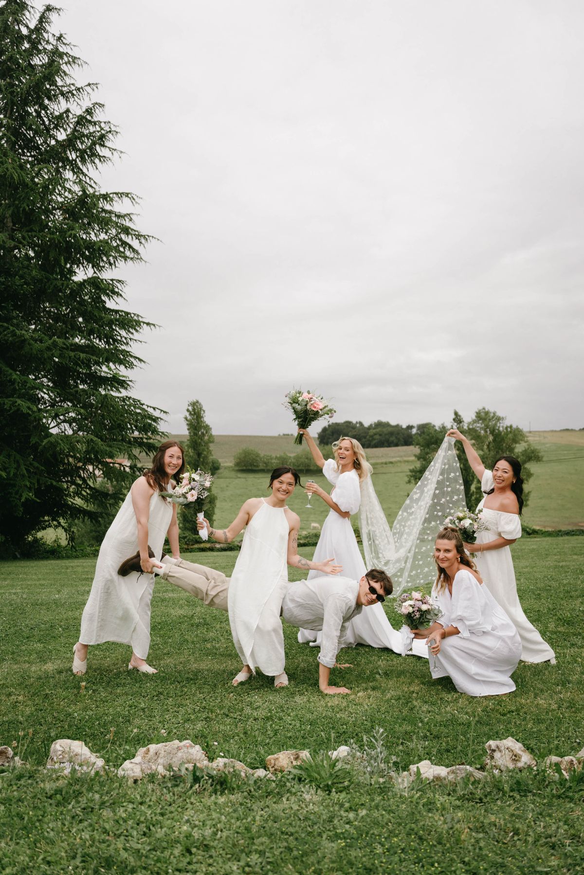Bridal party group portrait with bride and attendants in white dresses posing playfully in countryside field