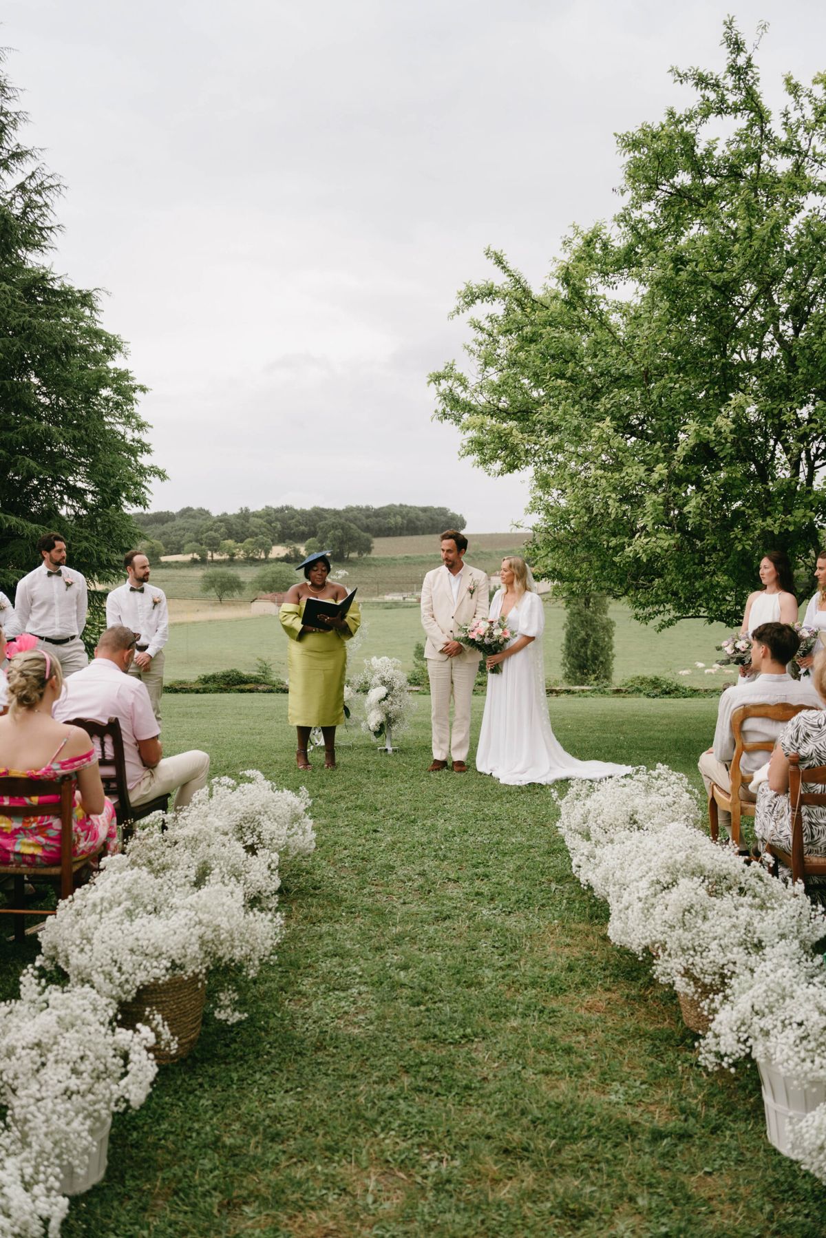 Outdoor ceremony on grass lawn with baby's breath lined aisle, couple at altar with countryside backdrop