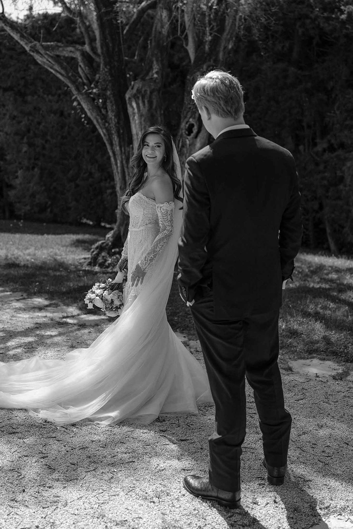Black and white first look with smiling bride in off-shoulder lace gown facing groom on gravel path