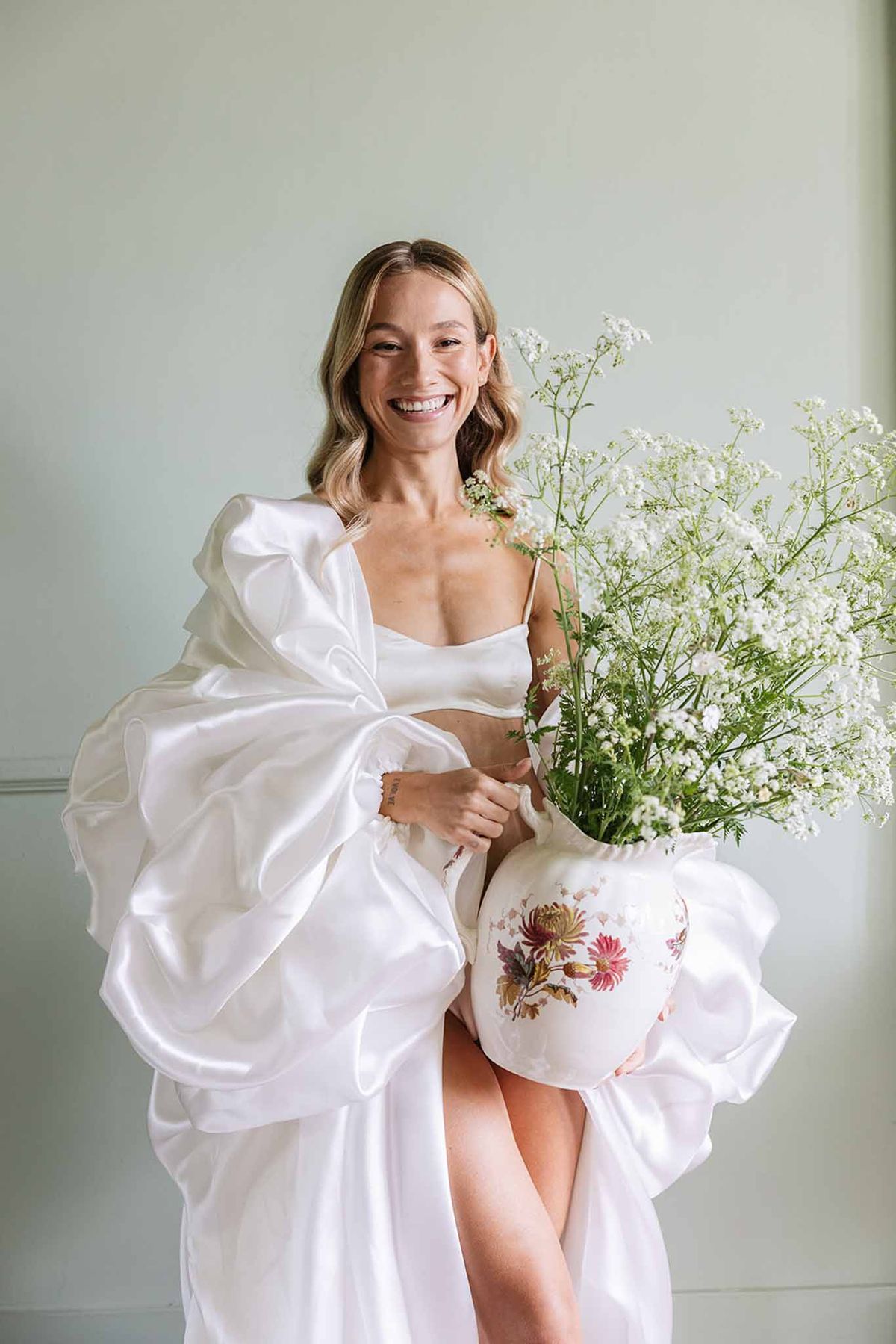 Bride in sculptural white gown holding vintage floral pitcher against pale green backdrop