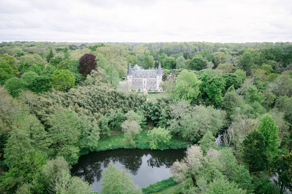 Aerial view of grey stone chateau with pointed turrets set within wooded estate beside dark pond