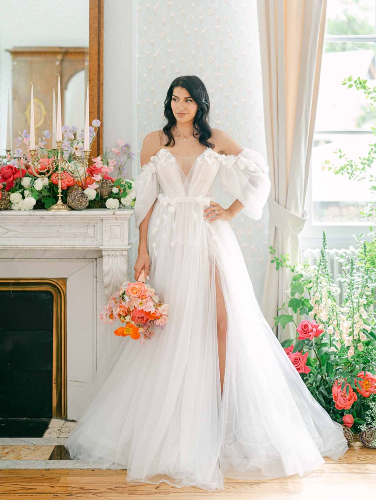 Bride in tulle V-neck gown with coral bouquet beside fireplace mantel of coral roses and pink tapers