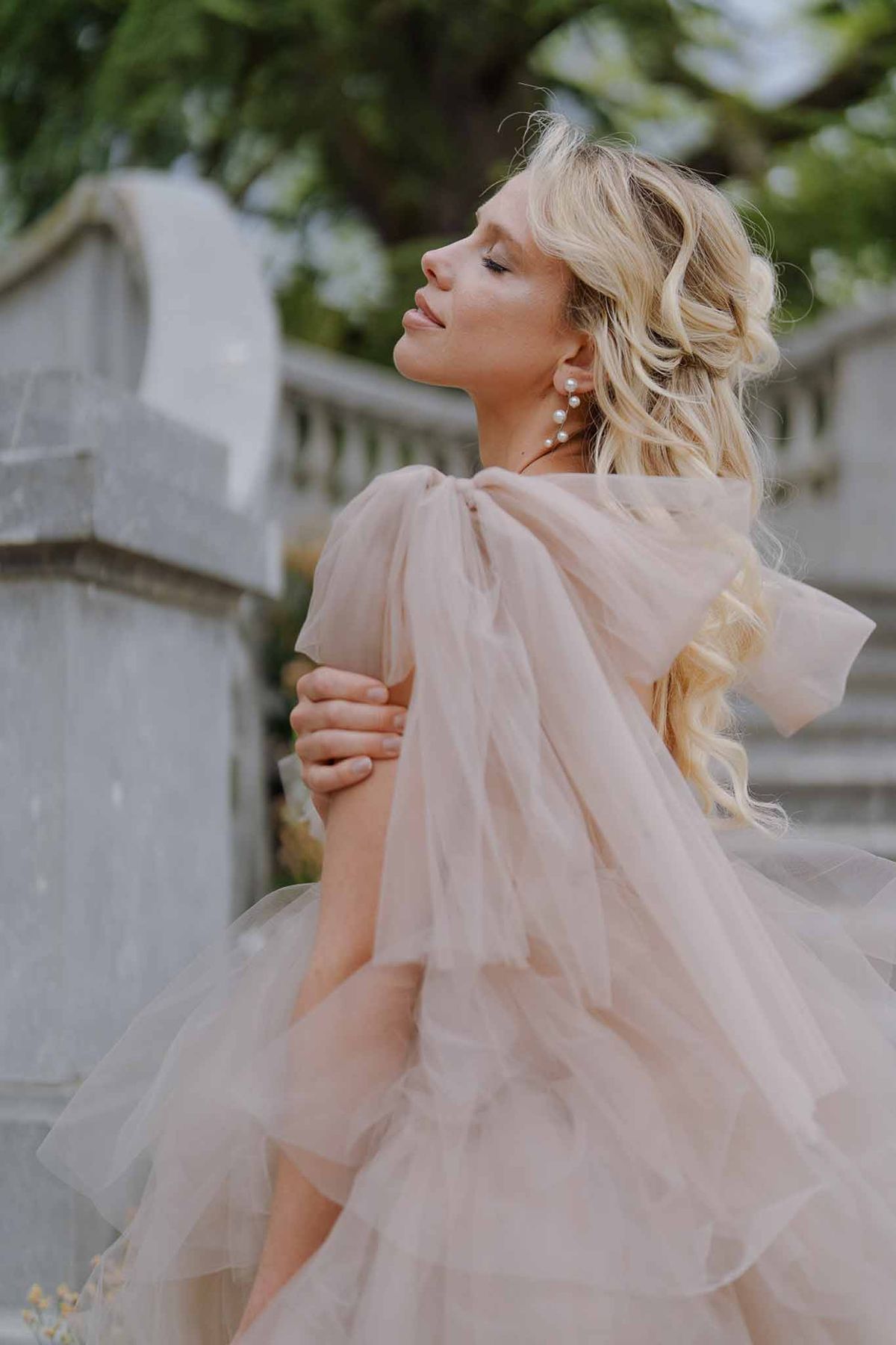 Bride with eyes closed wearing blush pink tulle off-shoulder gown and pearl drop earrings outdoors near stone balustrade