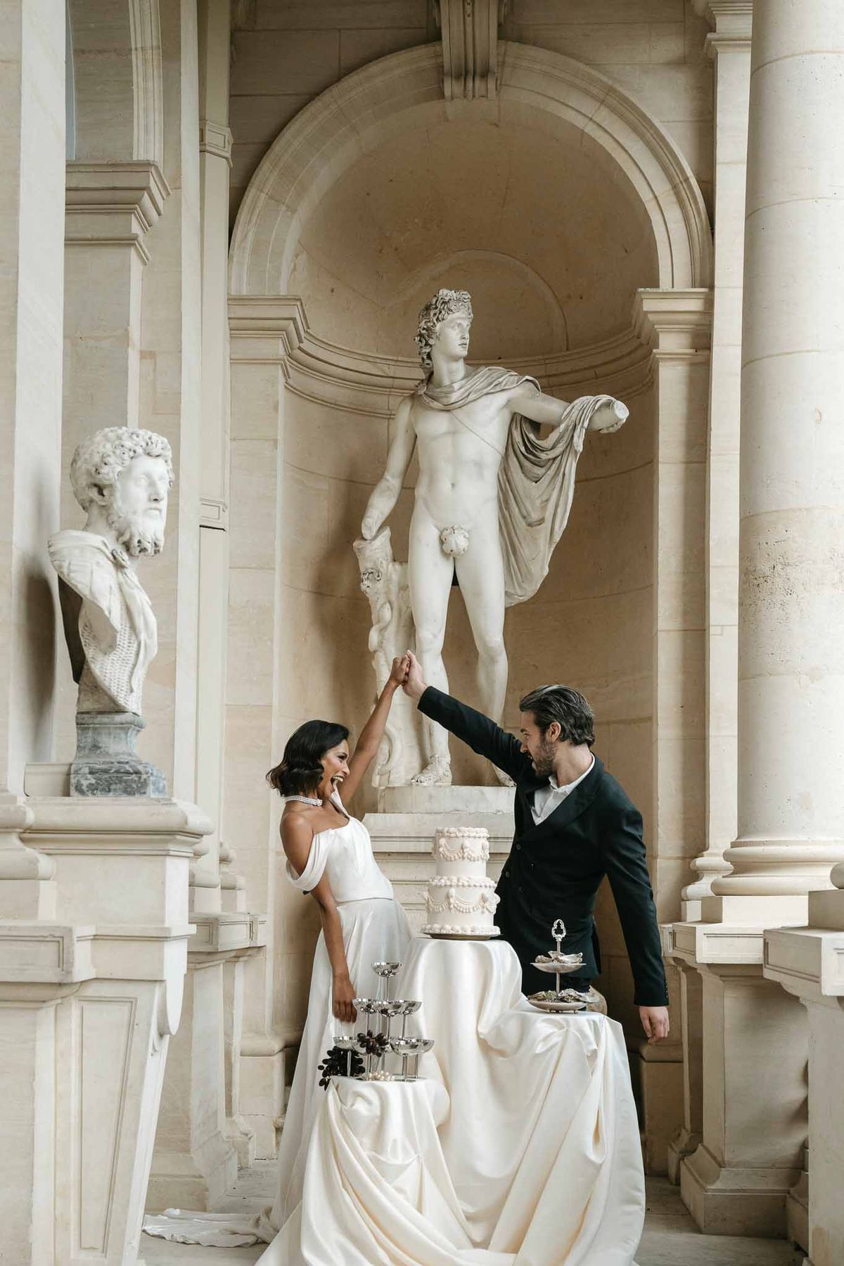 Bride and groom celebrating with wedding cake in neoclassical interior with columns and classical statues