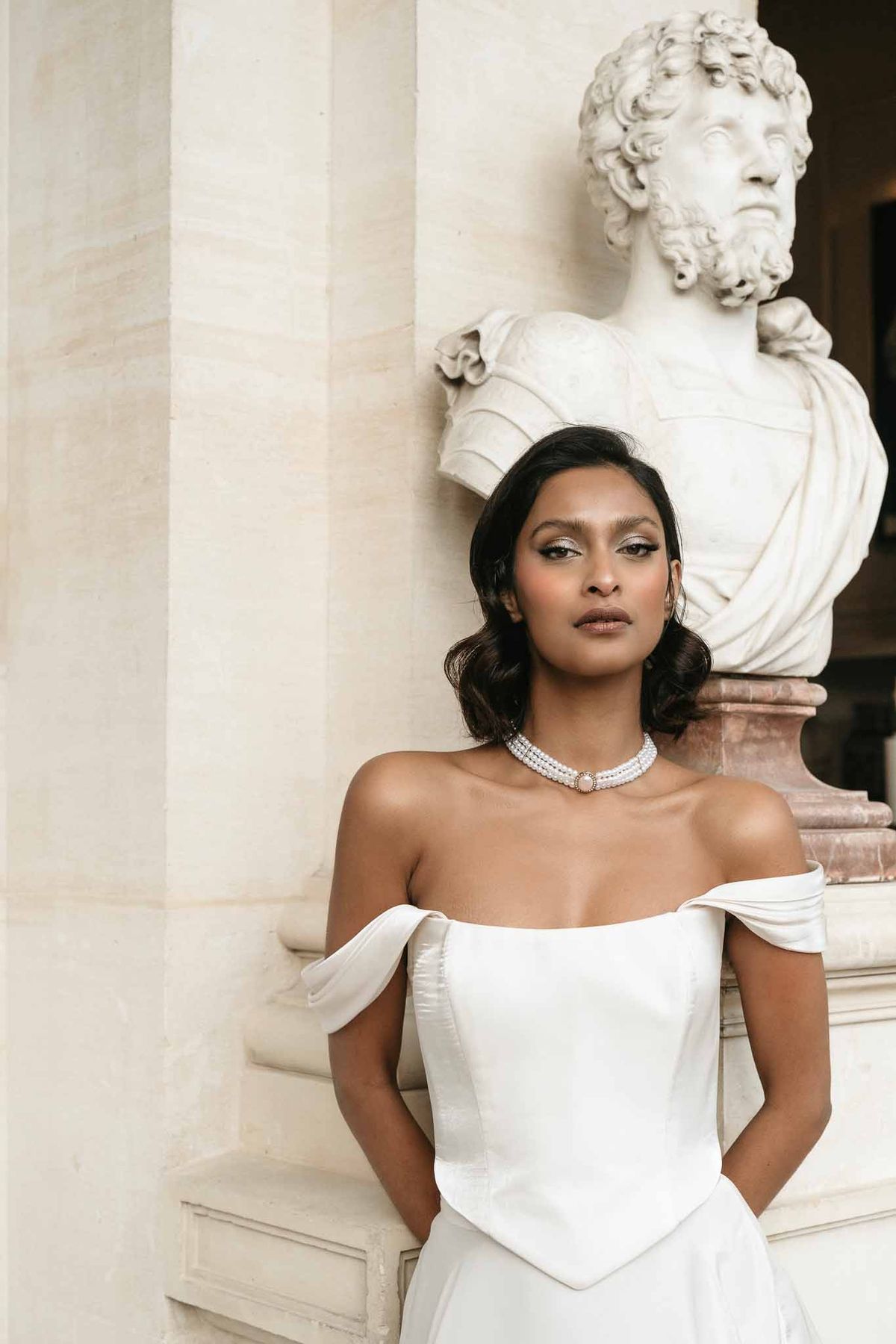 Bride in ivory gown with pearl necklace poses beside marble bust at classical venue with columns