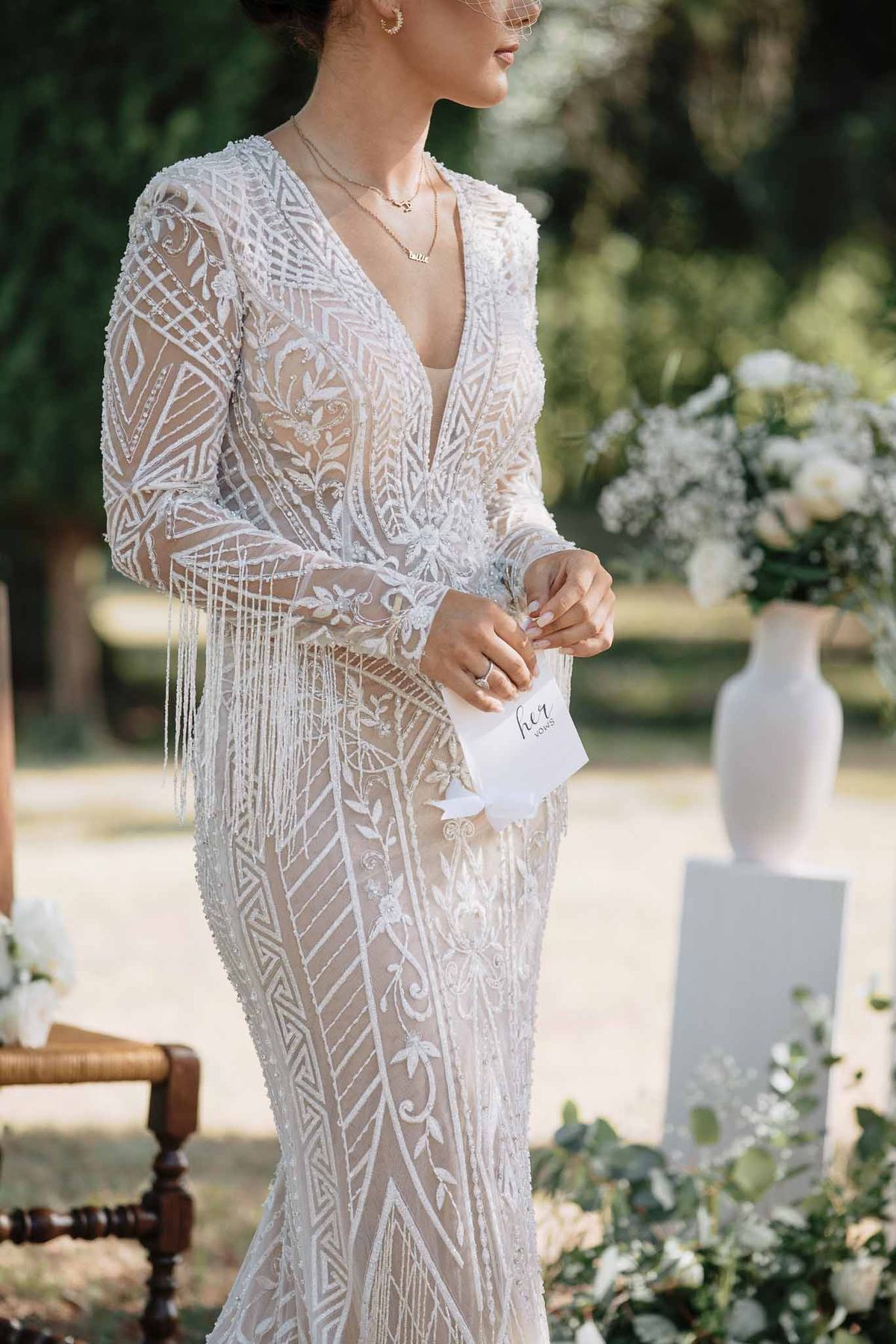 Close-up of bride holding vow booklet wearing beaded Art Deco long-sleeve gown with gold jewelry at outdoor ceremony