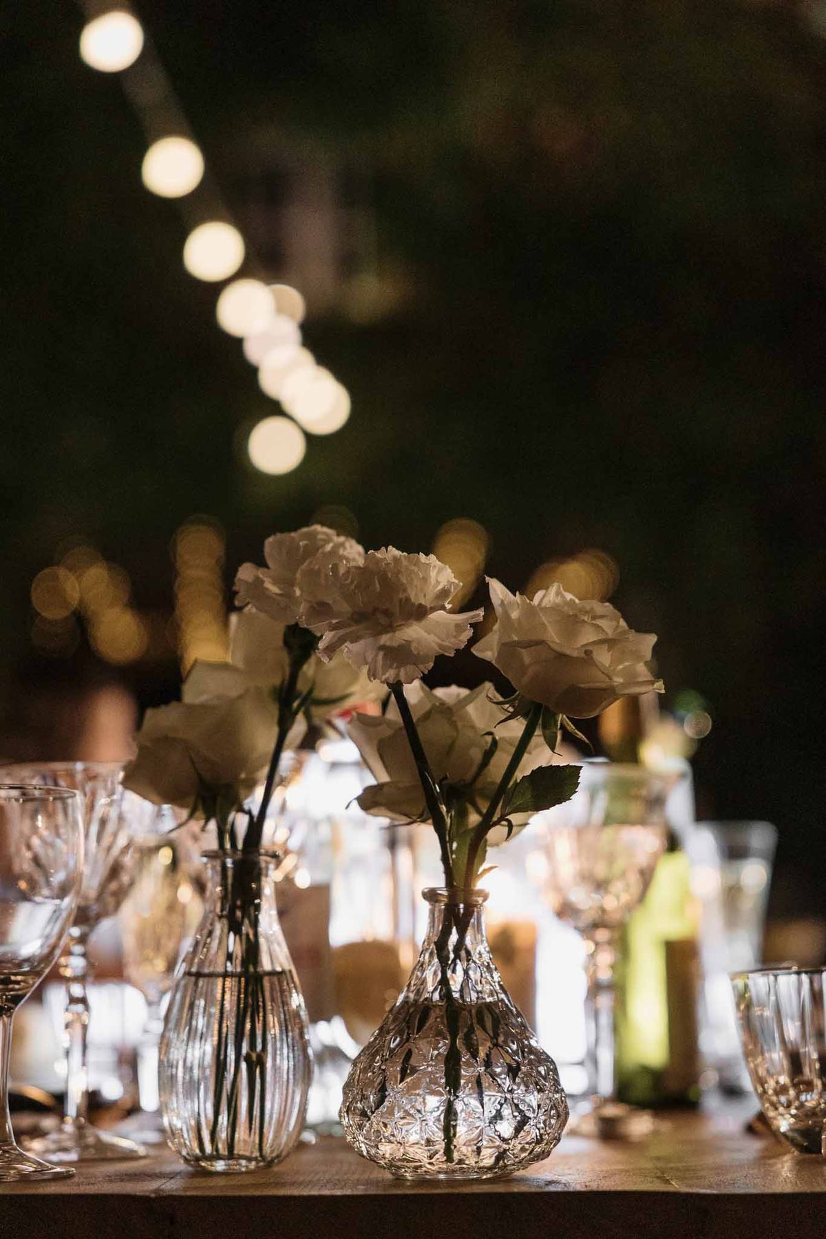 Cut-glass bud vases with white roses and peonies lit by candlelight with globe string light bokeh