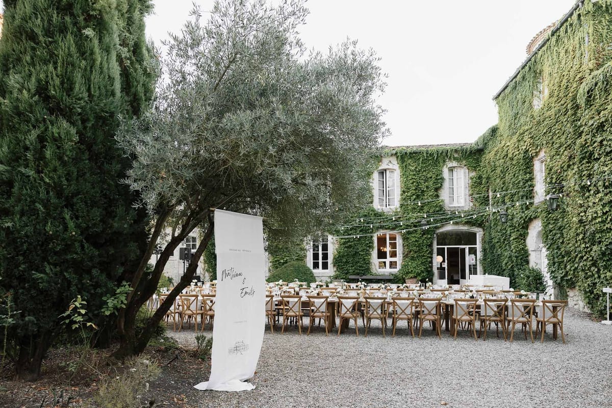Ivy-covered manor courtyard with long white tables, cross-back chairs, bistro lights, and calligraphy welcome sign