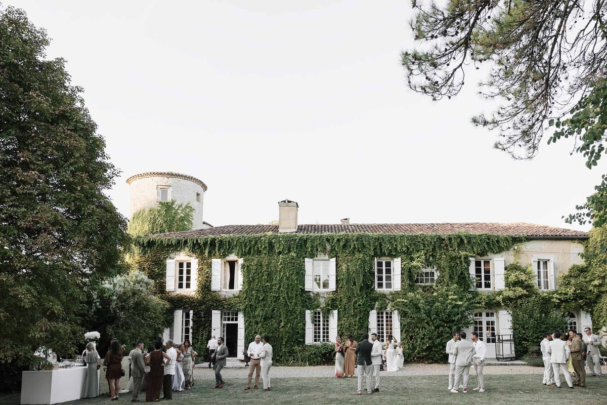 Thirty guests mingling on lawn before ivy-covered chateau with round tower at dusk cocktail hour