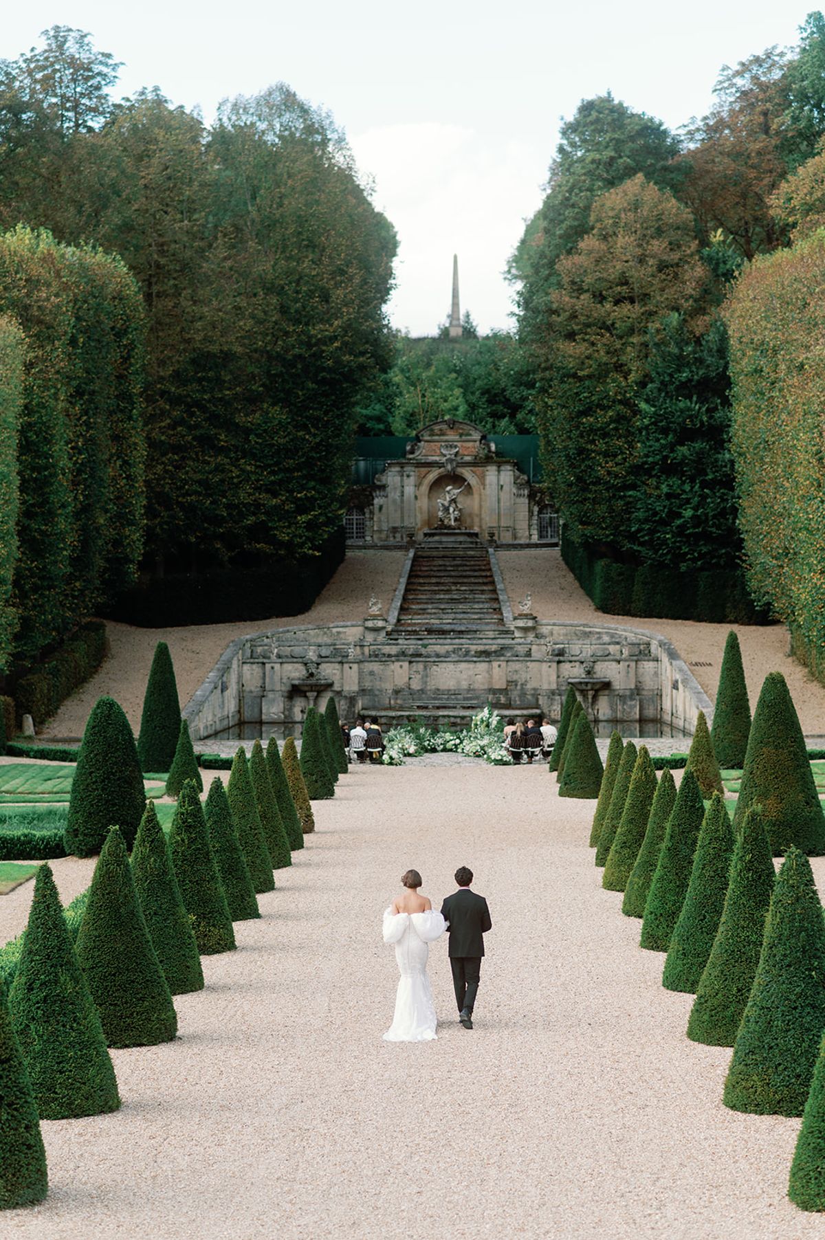 Couple walking topiary-lined allee toward ceremony at grand stone staircase in formal French garden