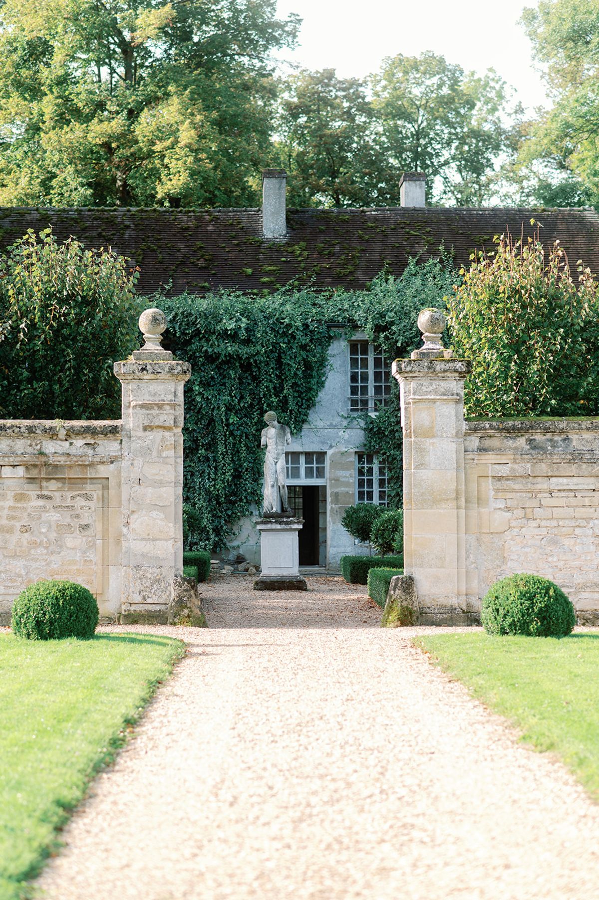 Stone gate pillars with boxwood spheres framing path to ivy-covered chateau with classical statue
