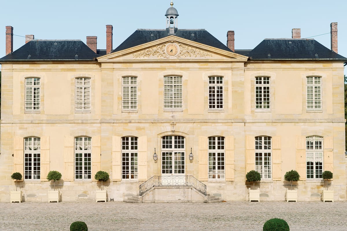 Golden limestone chateau with mansard roof, clock pediment, and topiary-lined cobblestone forecourt