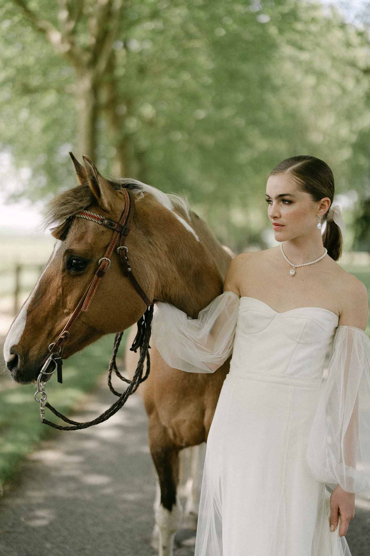 Bride in white gown posing with chestnut horse in tree-lined avenue for equestrian wedding portrait