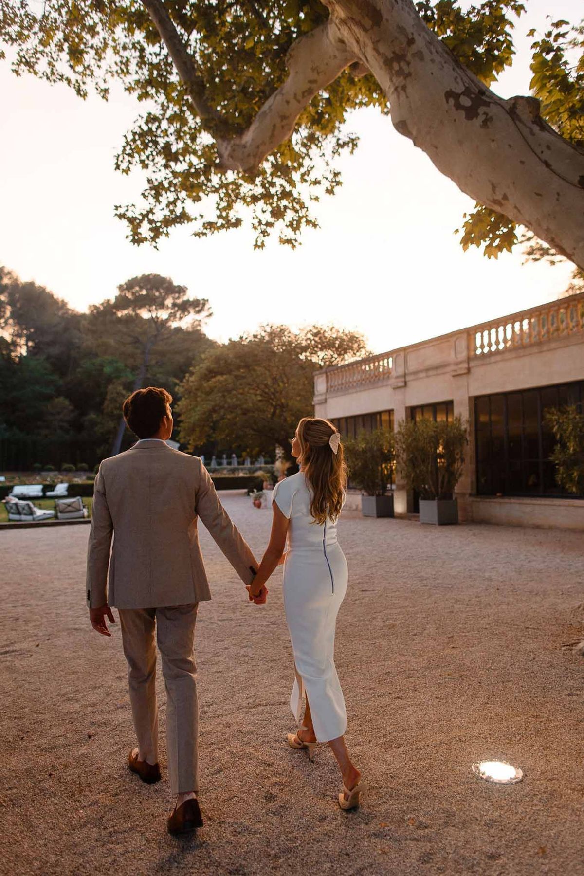 Bride and groom walking hand-in-hand across courtyard of classical stone mansion during golden hour