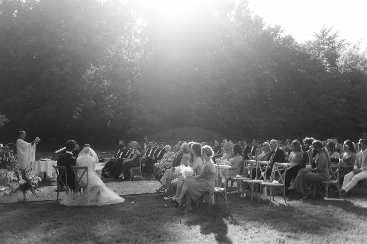 Outdoor wedding ceremony with guests in wooden chairs in tree-lined garden setting