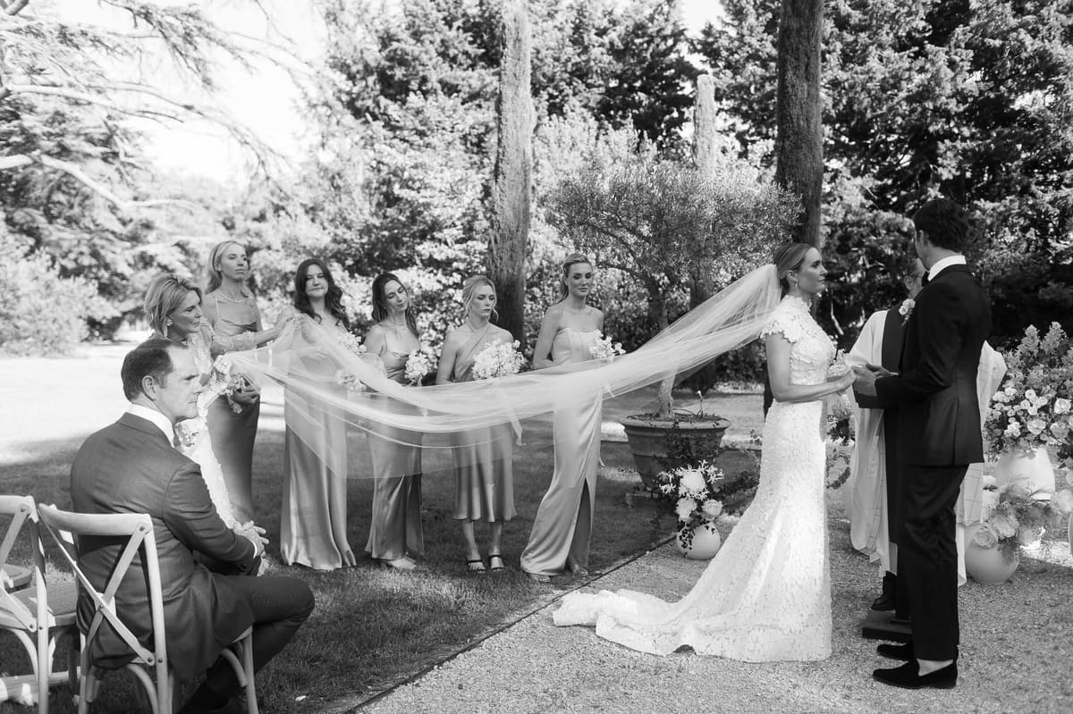 Outdoor wedding ceremony with bride's veil held by bridesmaids in garden setting with white floral arrangements