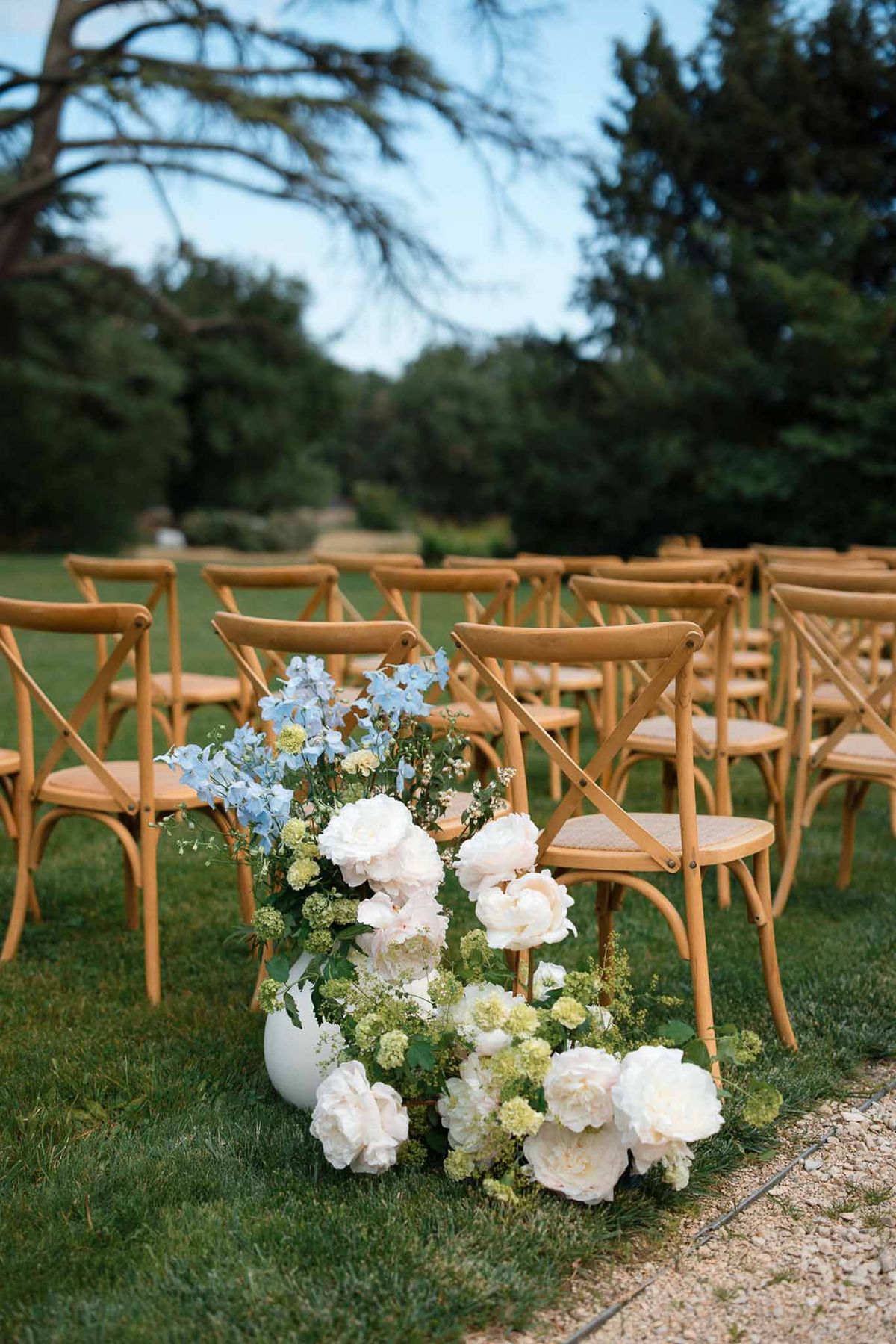 White and blue floral arrangement along ceremony aisle with wooden chairs in garden setting
