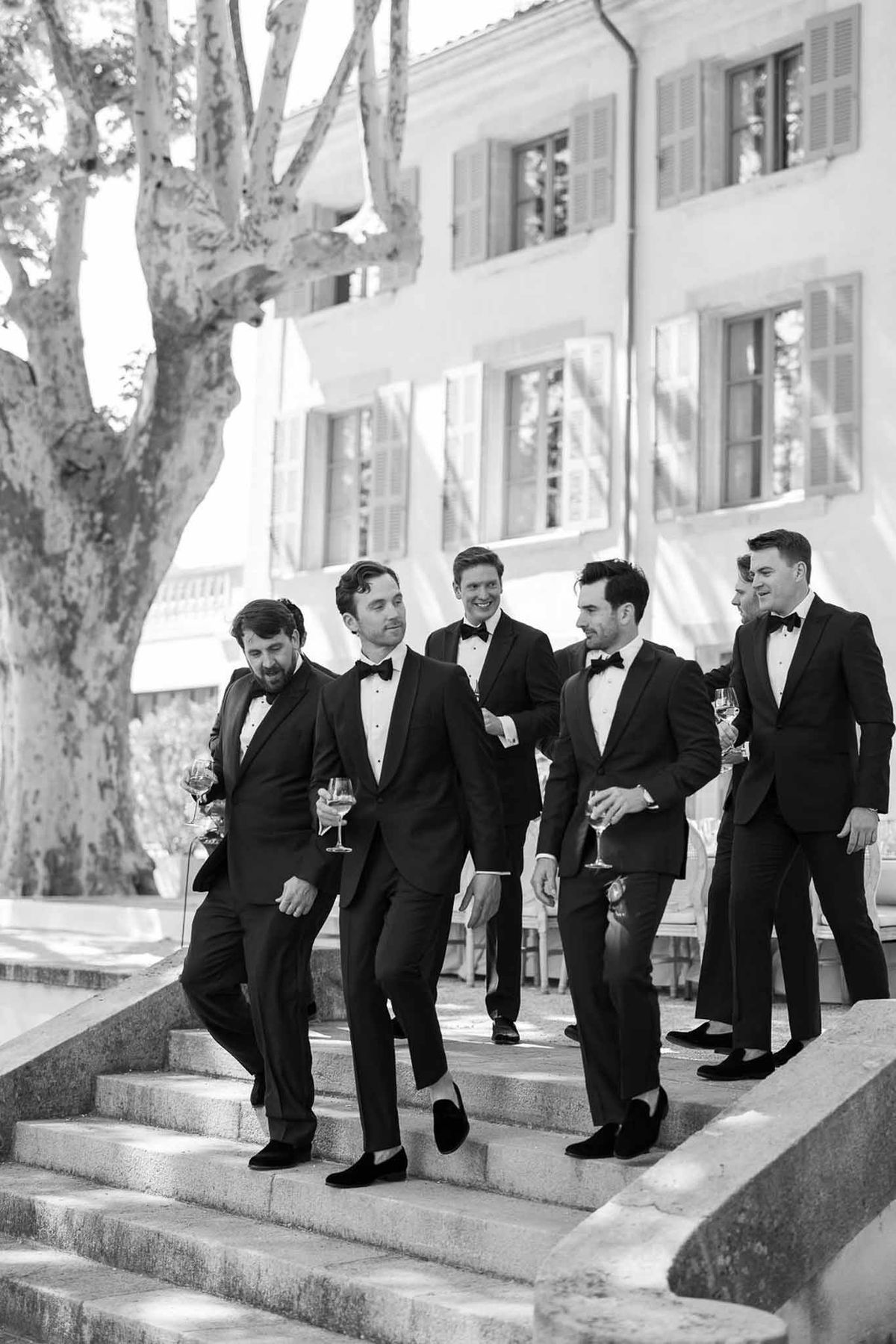 Groom and groomsmen in black tuxedos walking with champagne at outdoor wedding venue