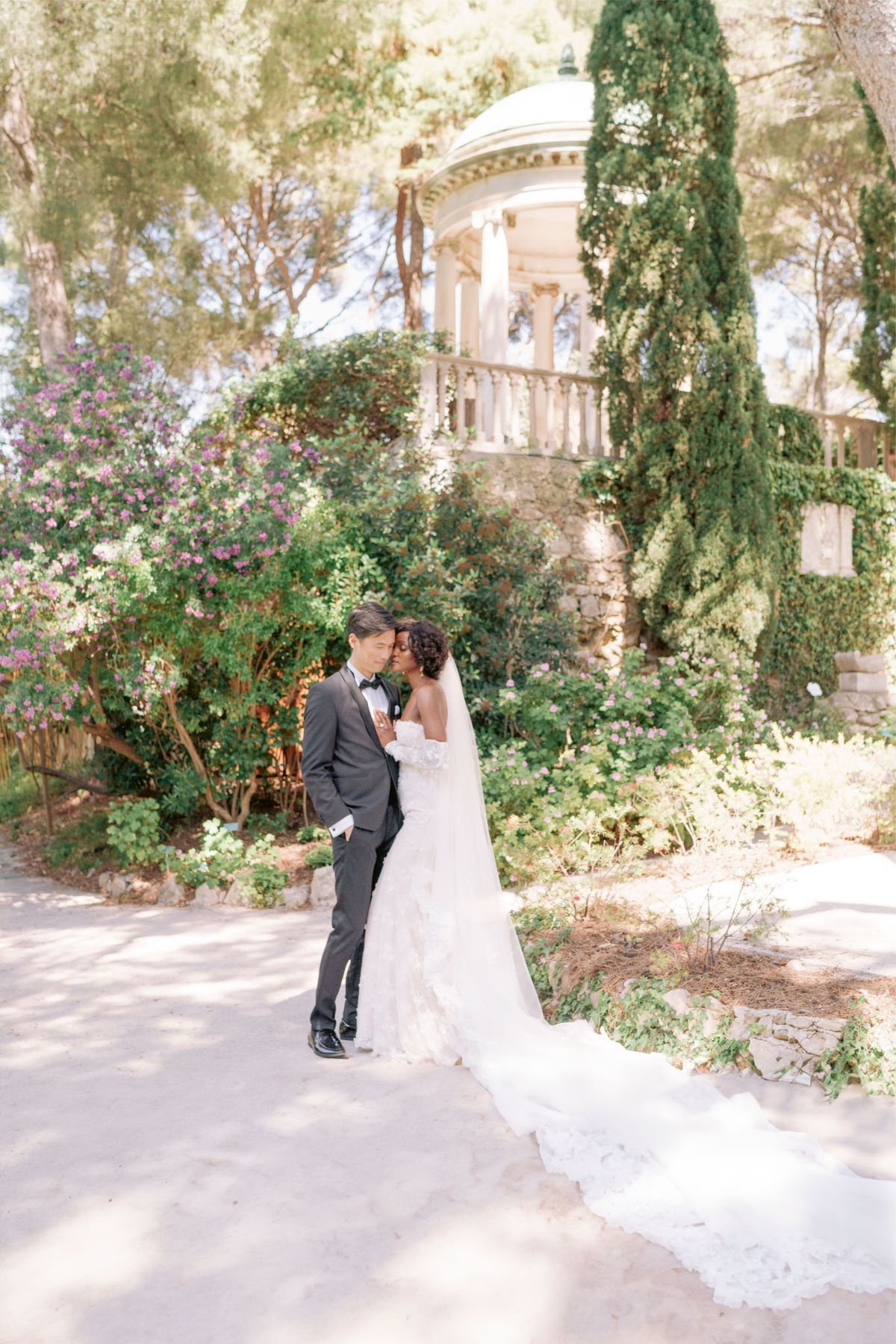 Couple embracing in formal garden with stone rotunda, bride in off-shoulder lace gown with cathedral veil