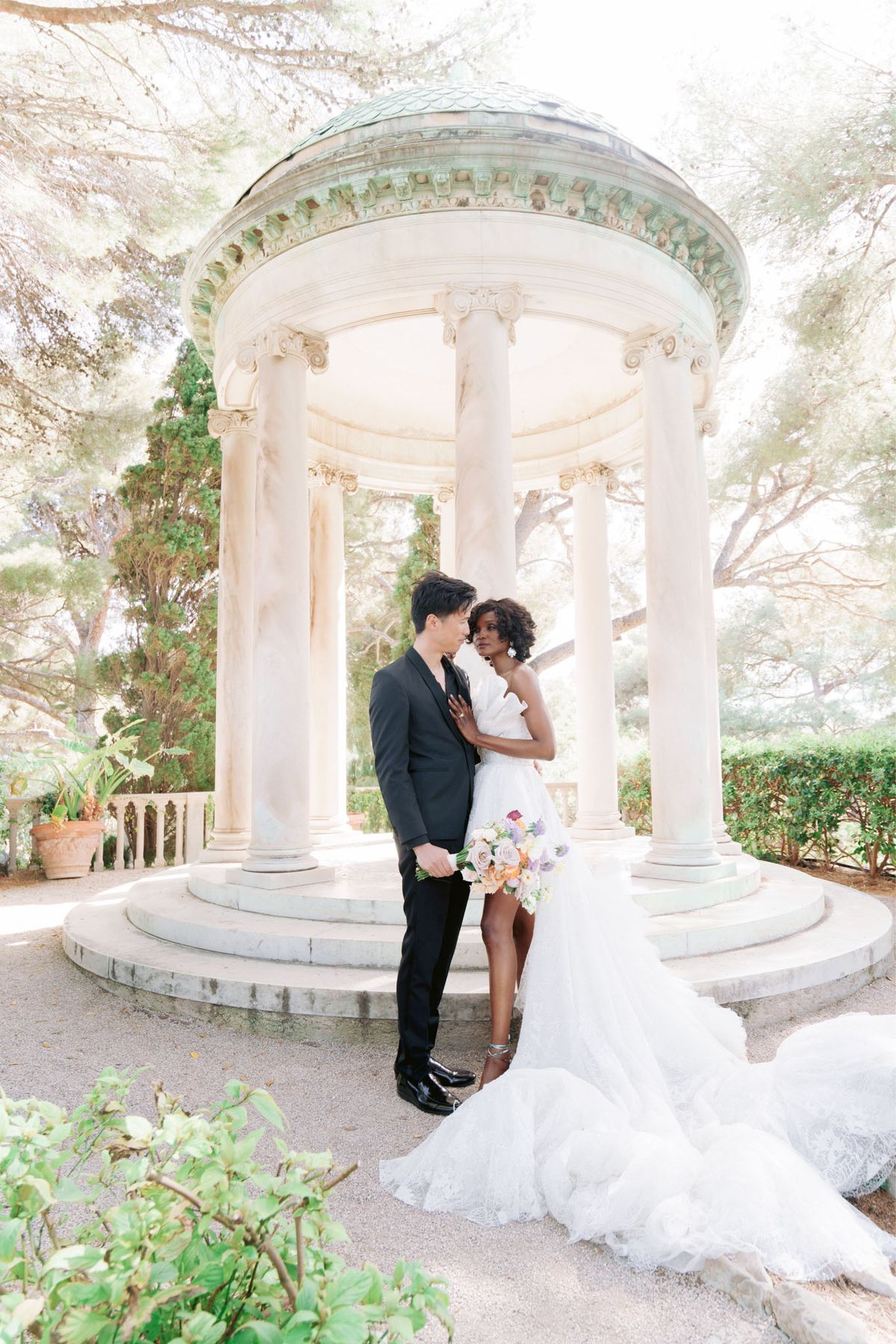 Couple before white marble Ionic rotunda with bride holding blush and lavender bouquet in formal garden