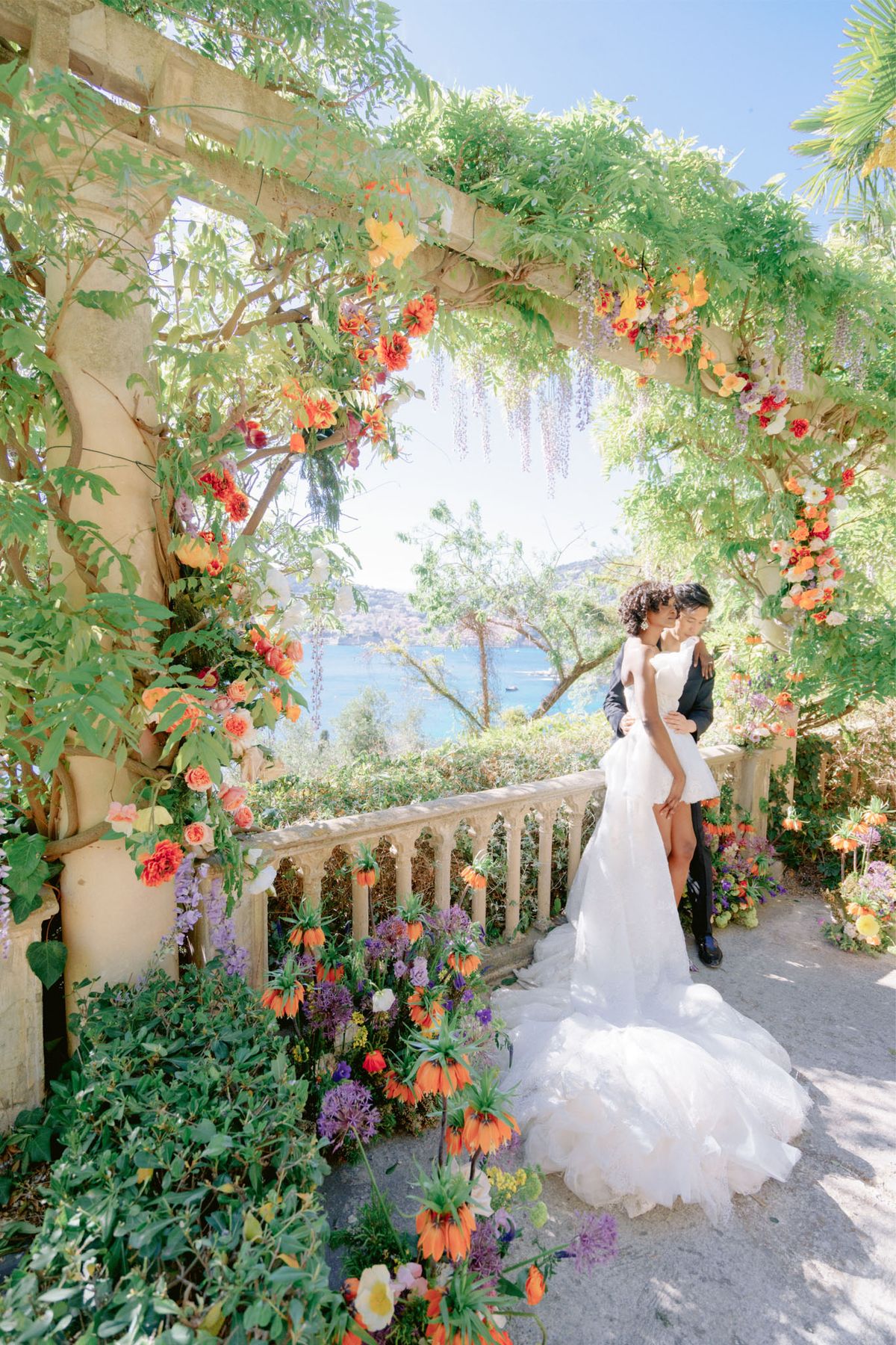Couple embracing under wisteria pergola with orange roses, purple allium, and coastal view beyond