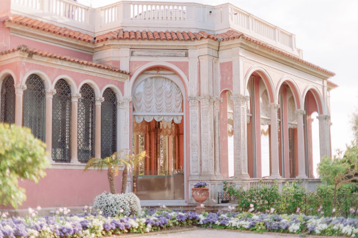 Coral-pink villa with white Corinthian colonnades, ivory drapes, and palm tree at arched entrance