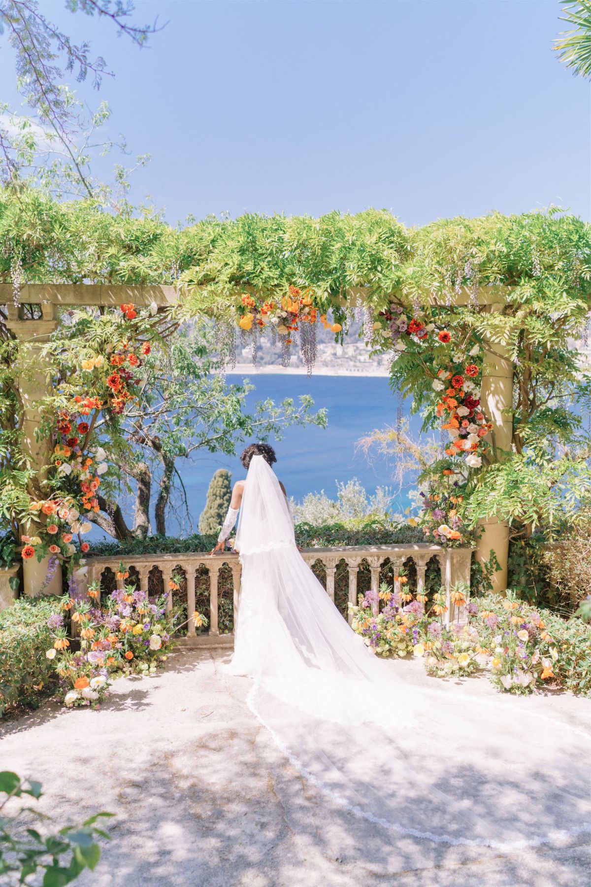 Bride from behind under wisteria pergola with orange and yellow floral columns overlooking blue Mediterranean water