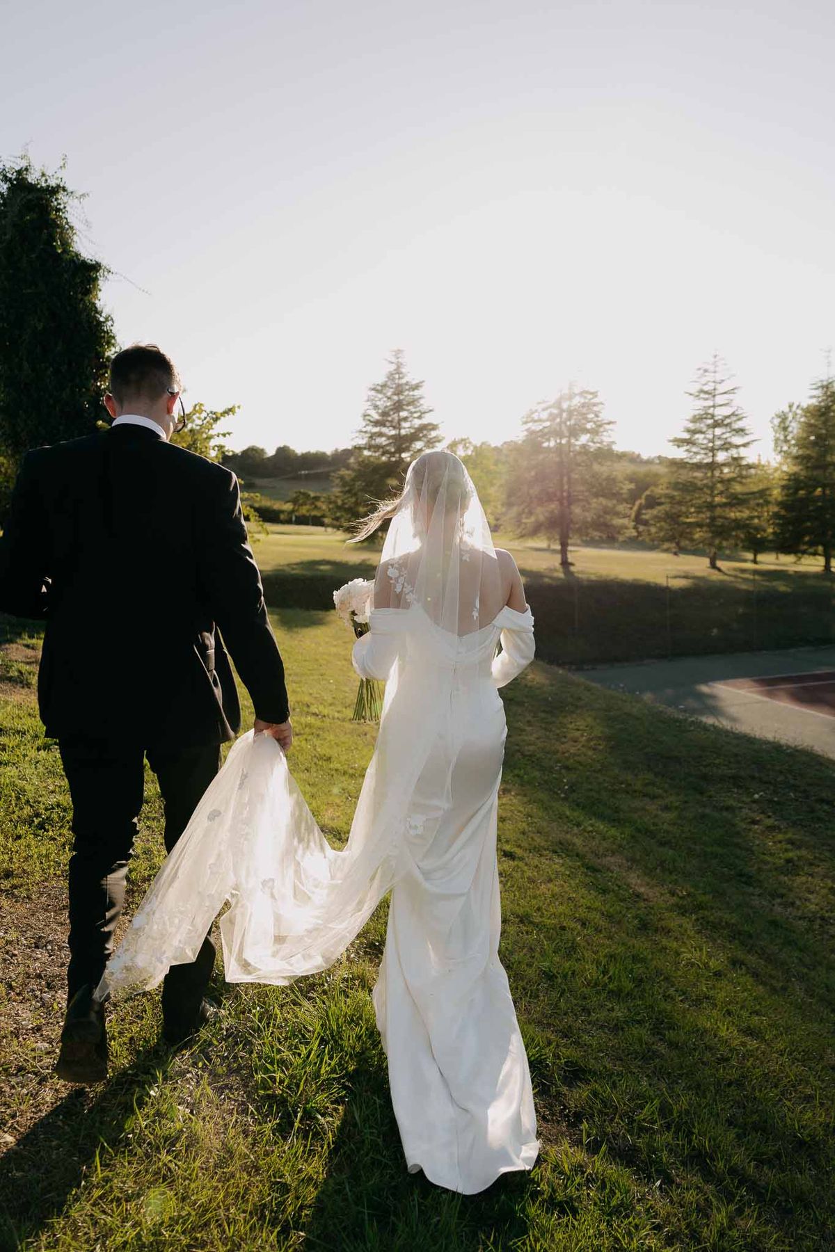 Couple walking across lawn at sunset with groom holding cathedral lace veil in golden backlight