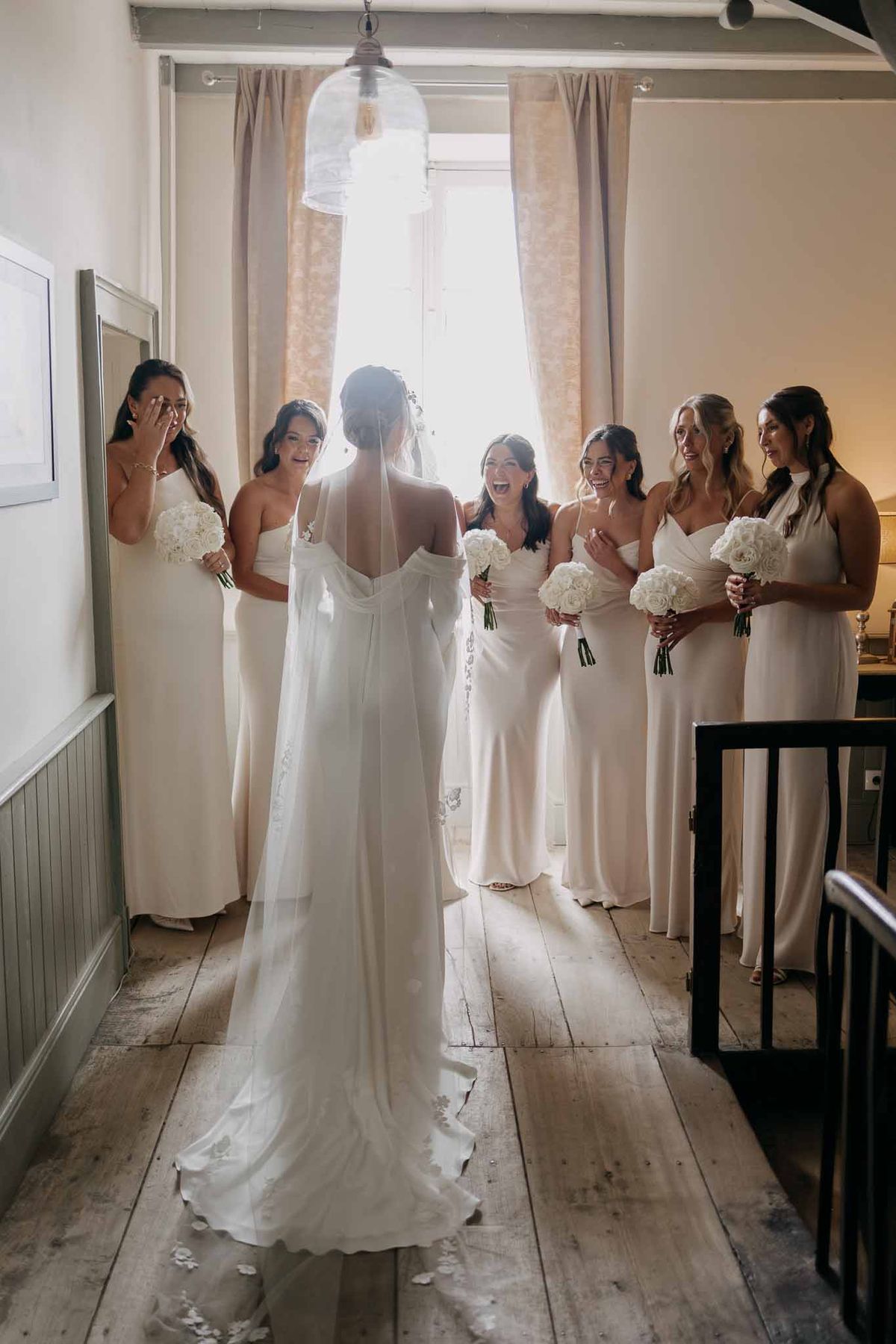Bride reveals dress to six bridesmaids in ivory dresses holding white rose bouquets with emotional reactions