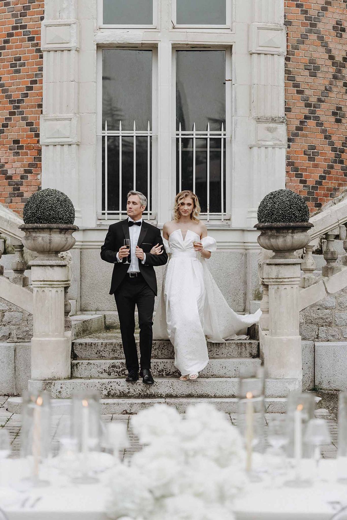 Bride and groom holding champagne flutes on stone staircase of red-brick manor with reception table in foreground