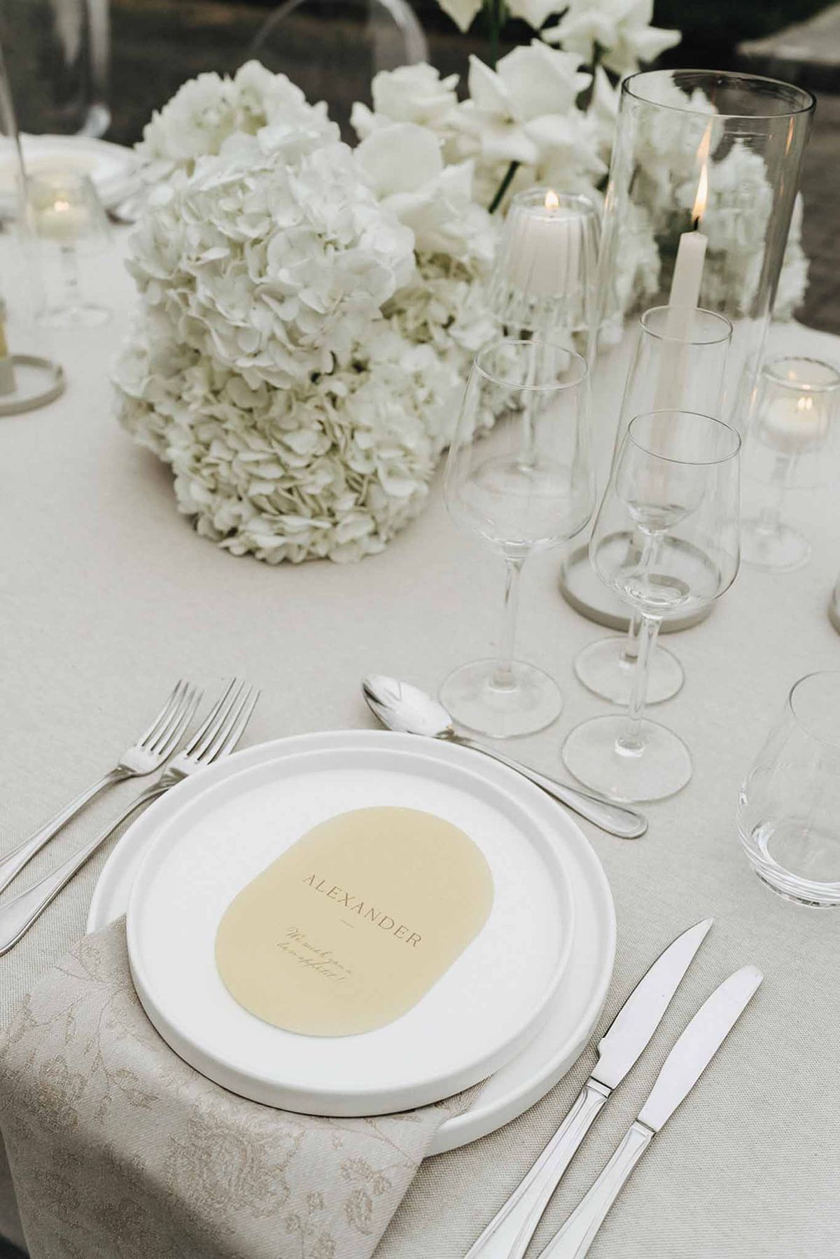Wedding reception place setting with white plates, gold script place card, hydrangea centerpiece, and crystal glassware