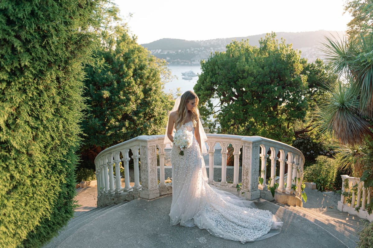 Bride on stone balustrade staircase in floral applique gown with veil, holding white rose bouquet, coastal backdrop