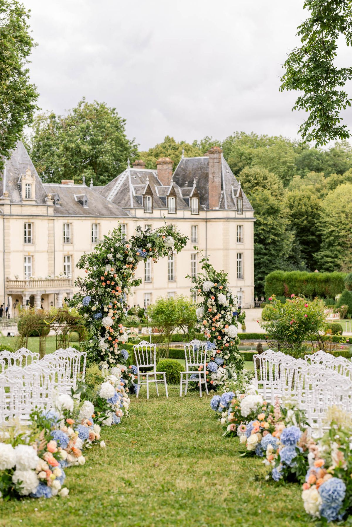 Blue hydrangea and coral rose floral arches frame altar chairs on lawn before mansard-roofed chateau