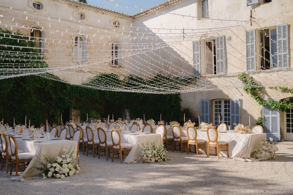 Long tables with blush ranunculus and fairy light canopy in ivy-walled chateau courtyard at evening