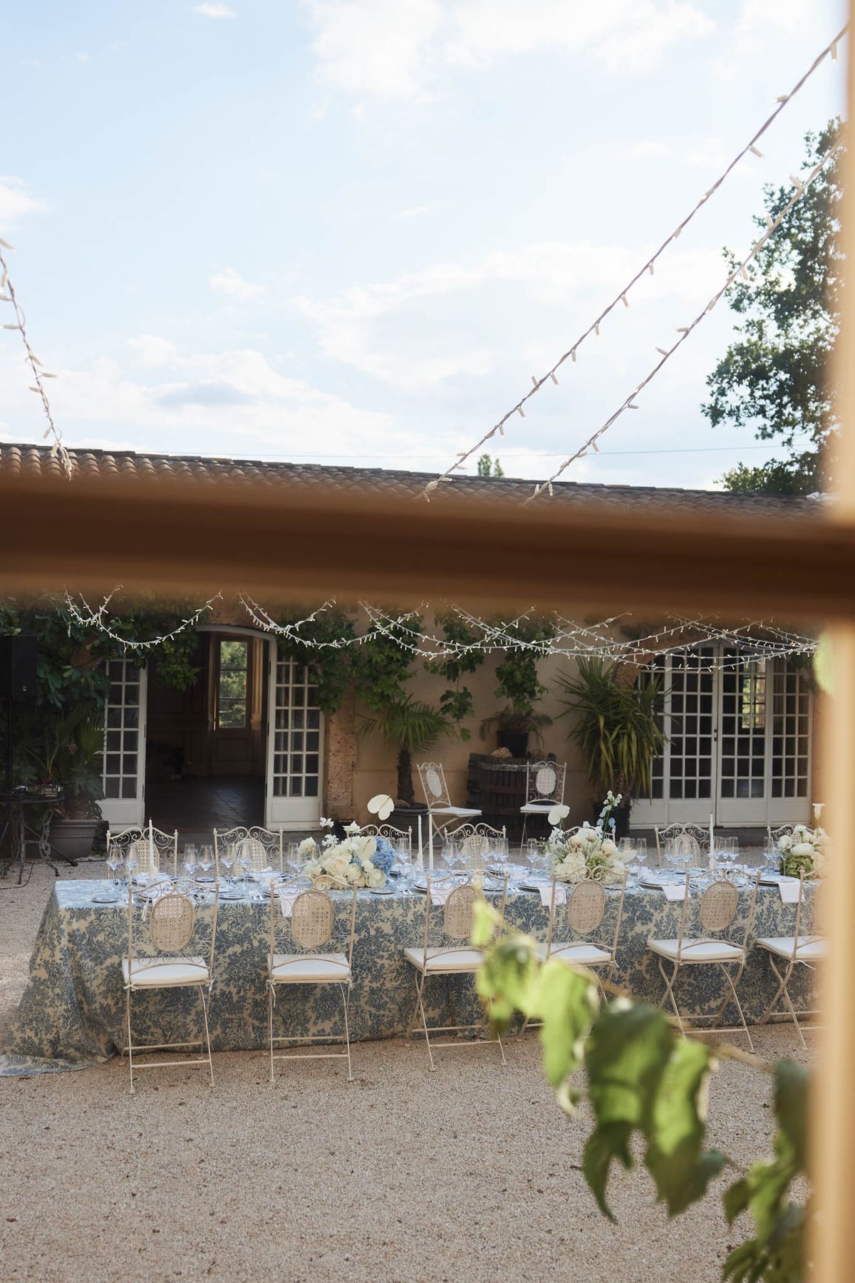Reception table with blue toile tablecloth, white roses, and gold bistro chairs in courtyard