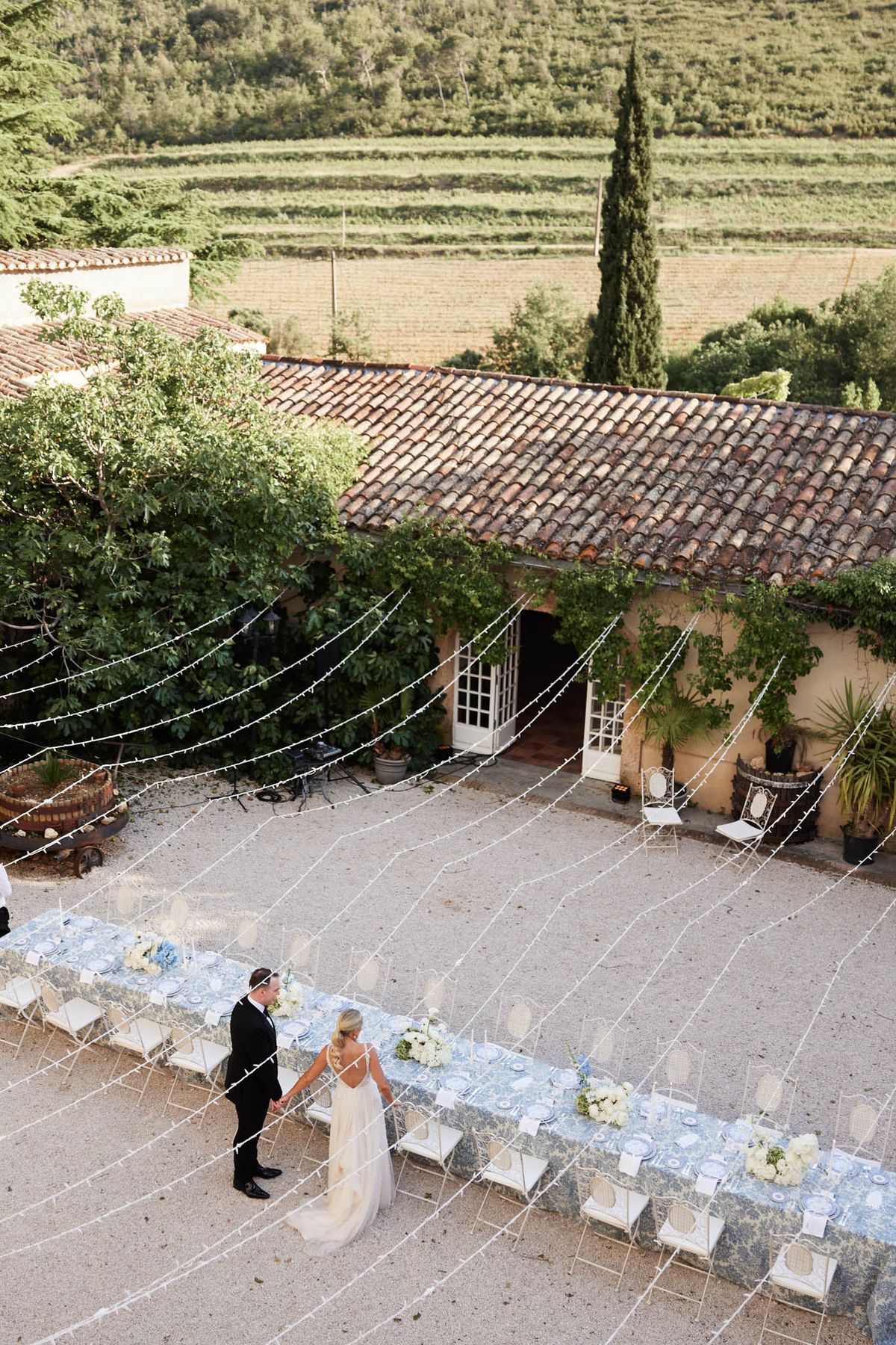 Aerial view of couple walking through courtyard reception with blue-and-white tablecloths and fairy light canopy