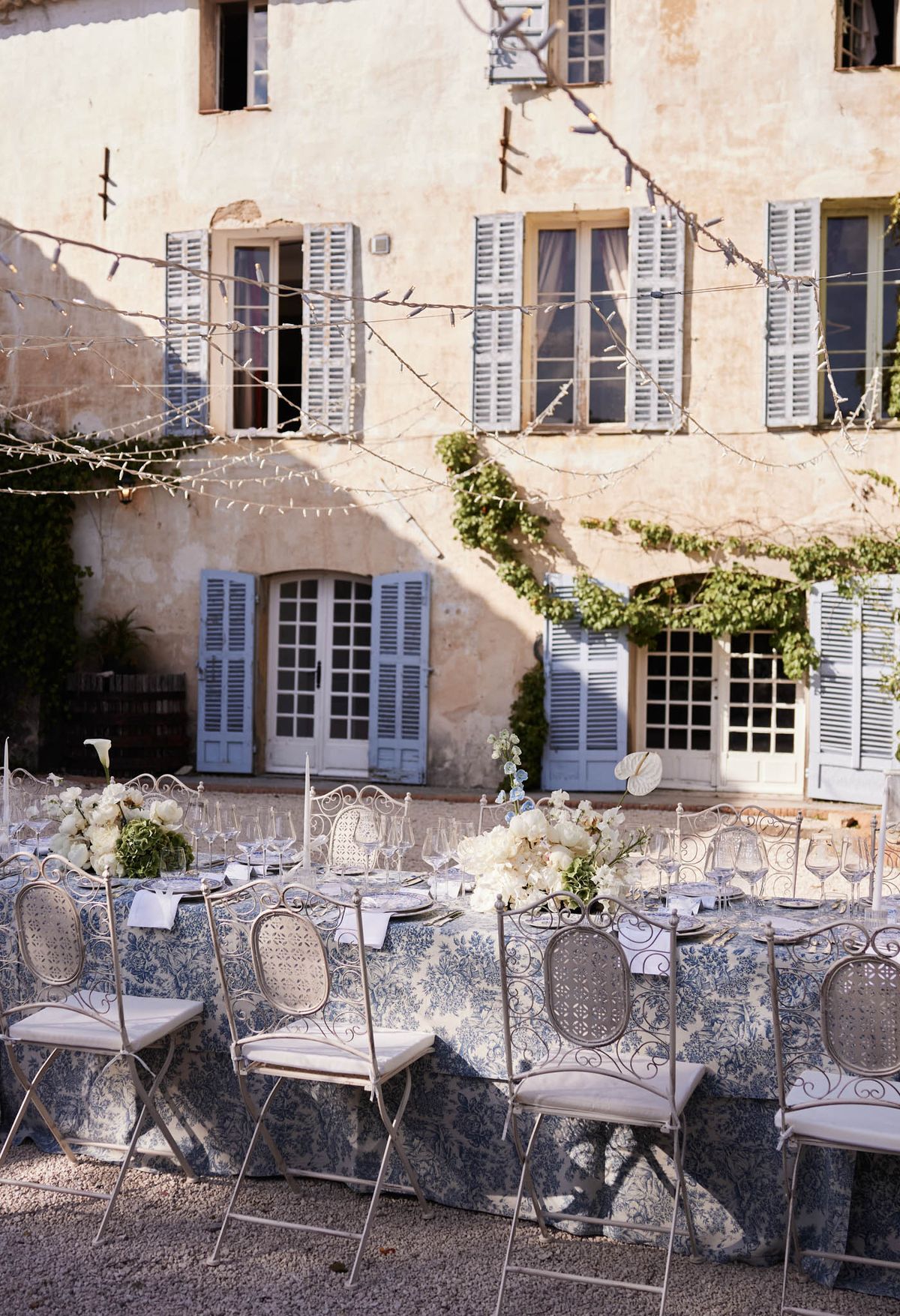 Toile de Jouy table with ivory peonies wrought-iron chairs and fairy lights before stone building