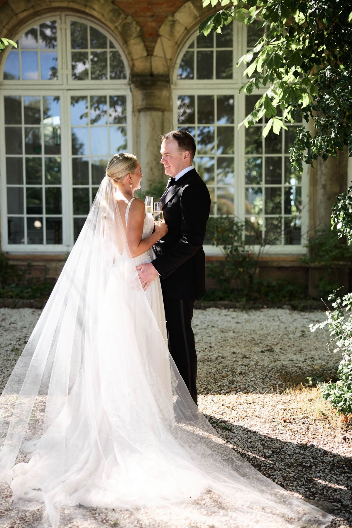 Bride in ivory tulle ballgown with cathedral veil and groom in tuxedo holding champagne on chateau gravel path