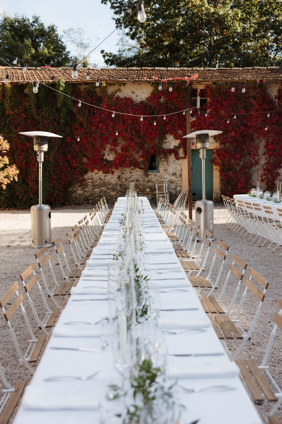 Long feasting table with olive-branch garland and Edison lights before vine-covered stone farmhouse