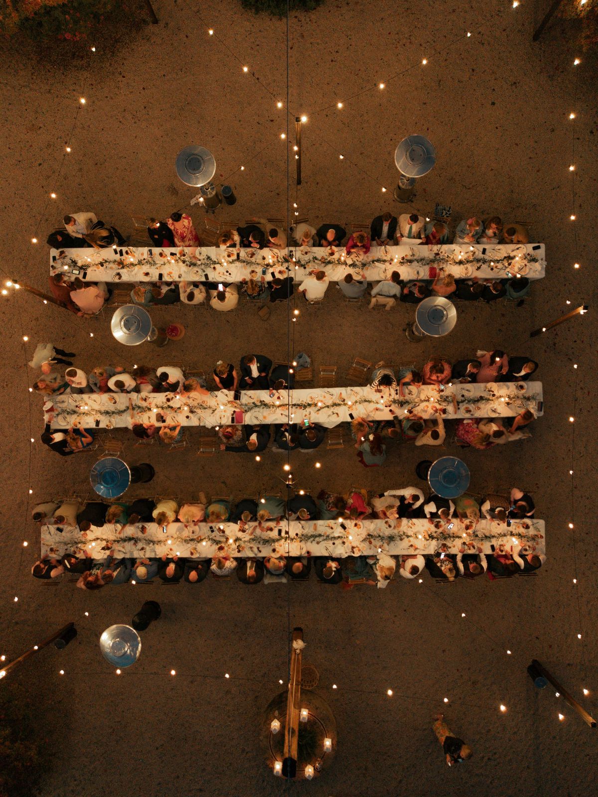 Aerial night view of three long candlelit tables with greenery runners and fairy lights seating one hundred guests